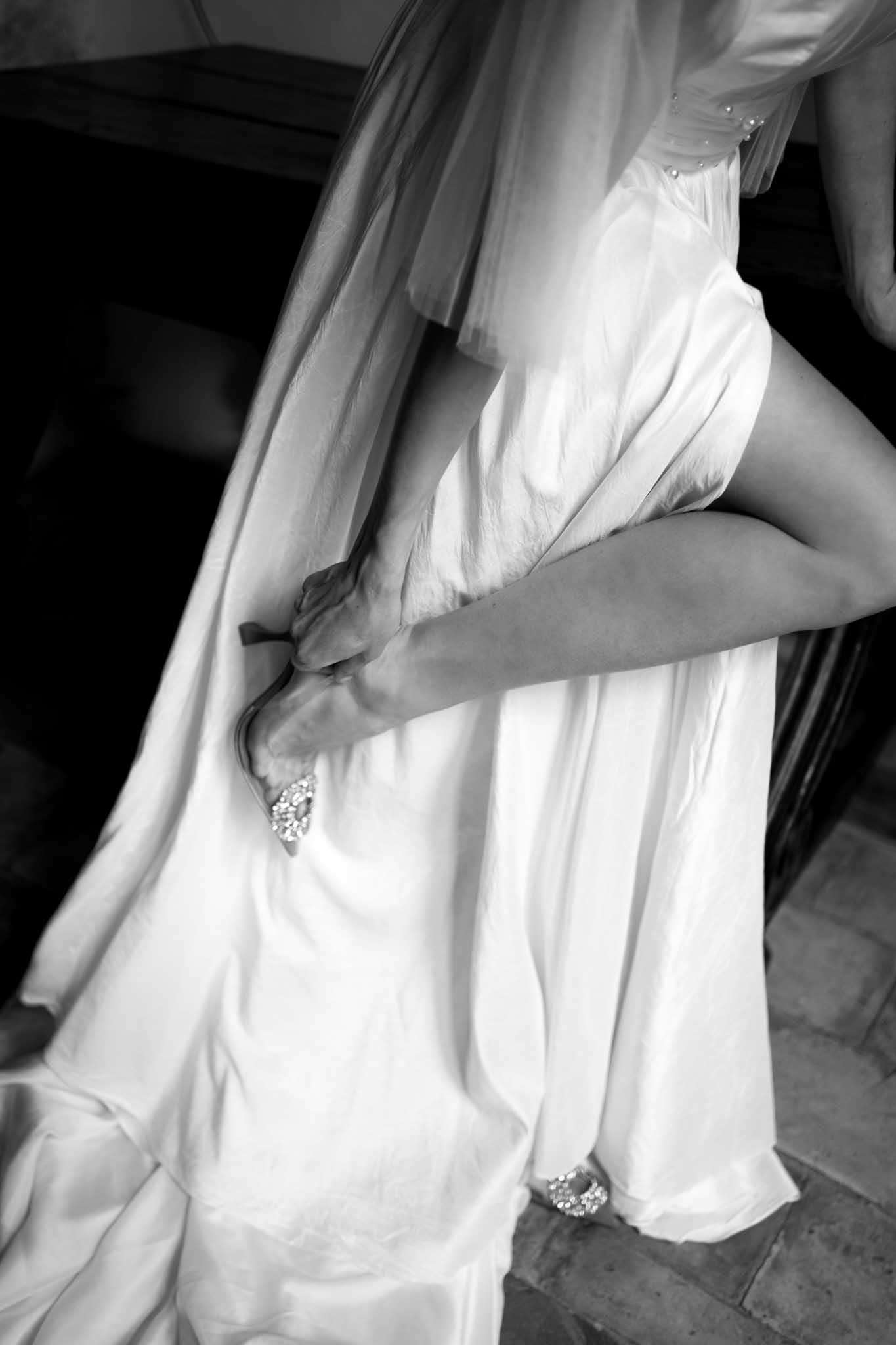 Black and white close-up of bride lifting satin gown skirt to reveal crystal-embellished heels with veil draping alongside