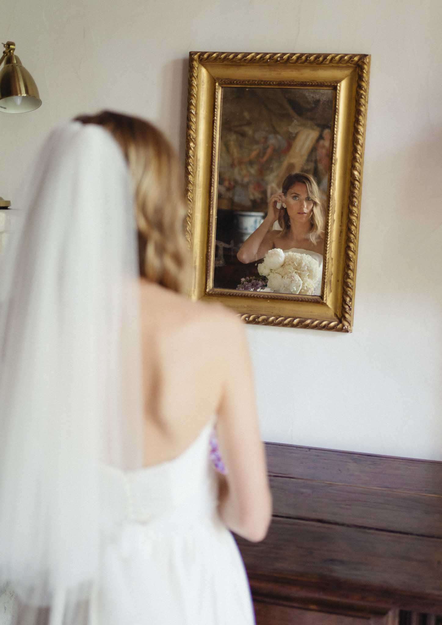 A bride is captured getting ready indoors, adjusting her earring while looking into an ornate gold-framed mirror mounted on a pale wall. The shot is composed from behind the bride, with her white veil and low-back ivory strapless gown in soft focus in the foreground, while her reflection — sharp and centered — shows her face, drop earrings, and a bouquet of white peonies held at her side. The room features a dark wood console table and a brass wall sconce, suggesting a classic, antique-furnished interior such as a château or manor house. The overall styling is classic and refined, with a soft, natural light palette.