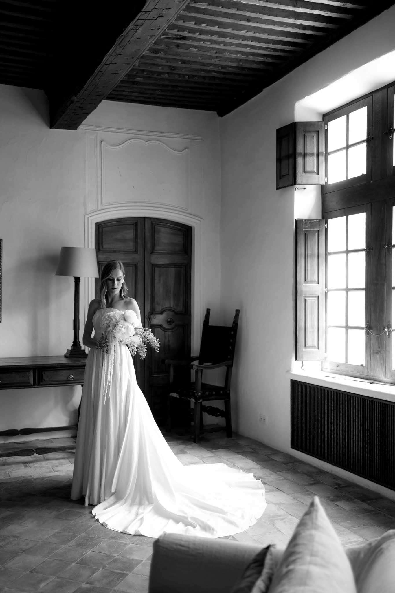 Black and white bride in strapless gown with trailing bouquet in chateau room with beamed ceiling and shutters