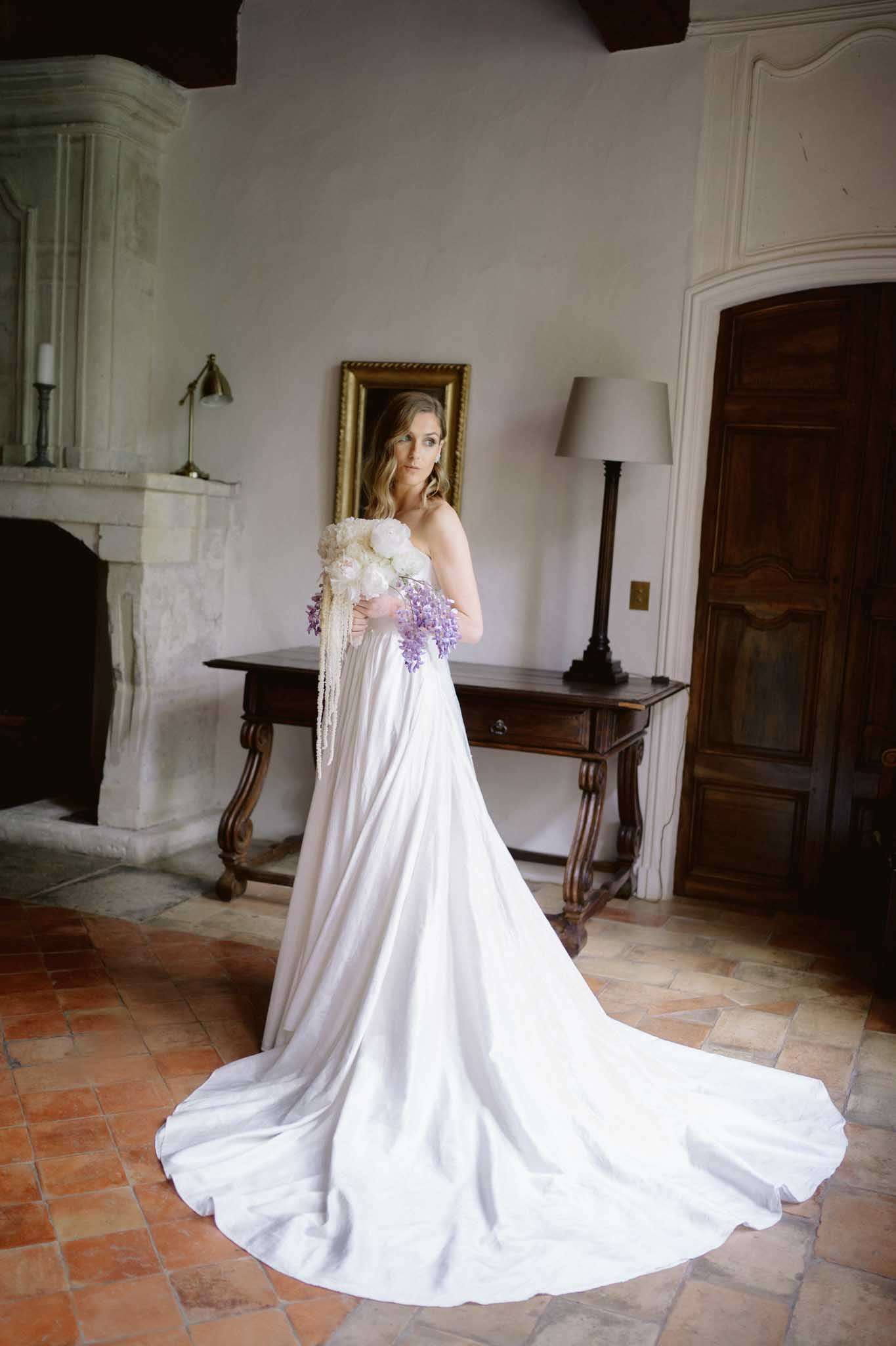 A bridal portrait taken indoors in what appears to be a French château interior, featuring a single bride standing on terracotta tile flooring. She wears a strapless ivory satin gown with a long cathedral-length train pooling on the floor, and holds a cascading bouquet of white peonies, purple wisteria, and trailing ivory amaranthus. The room features a large stone fireplace, exposed dark wood ceiling beams, white plaster walls, a dark antique wooden console table, a gold-framed mirror, and a table lamp, giving the space a classic French country interior feel. The shot is a full-length portrait with the bride facing the camera at a slight angle.
