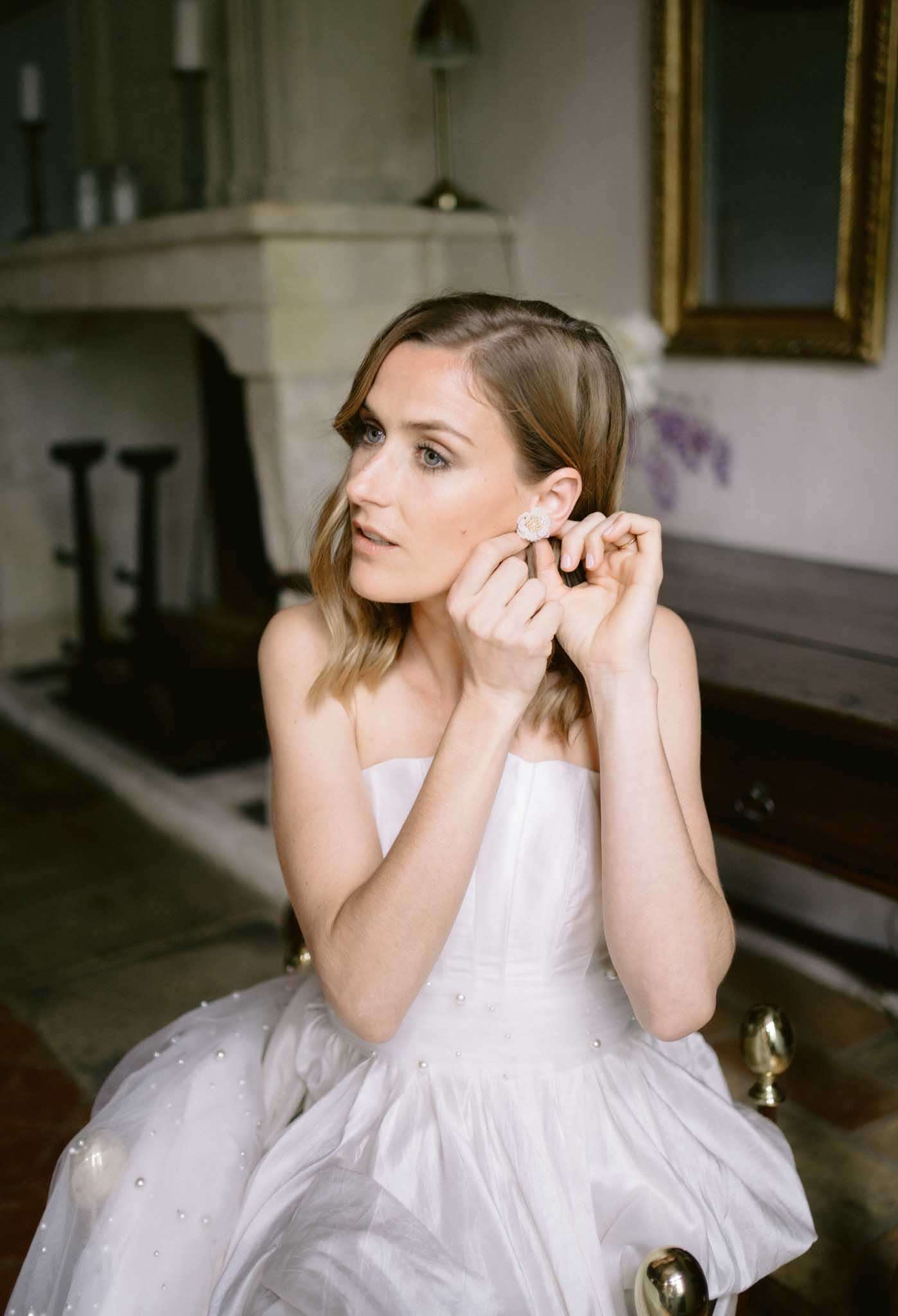 Bride fastening crystal earring wearing strapless pearl-embellished ballgown in chateau room with fireplace