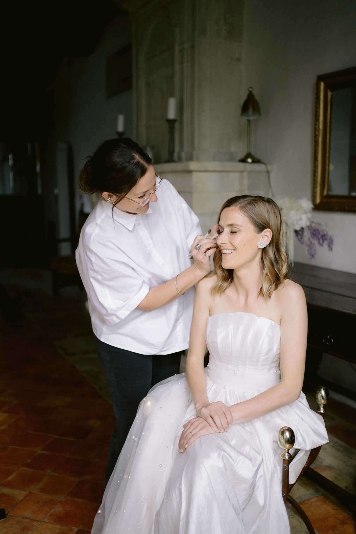 Makeup artist applying makeup to smiling bride in white gown inside chateau room with stone fireplace