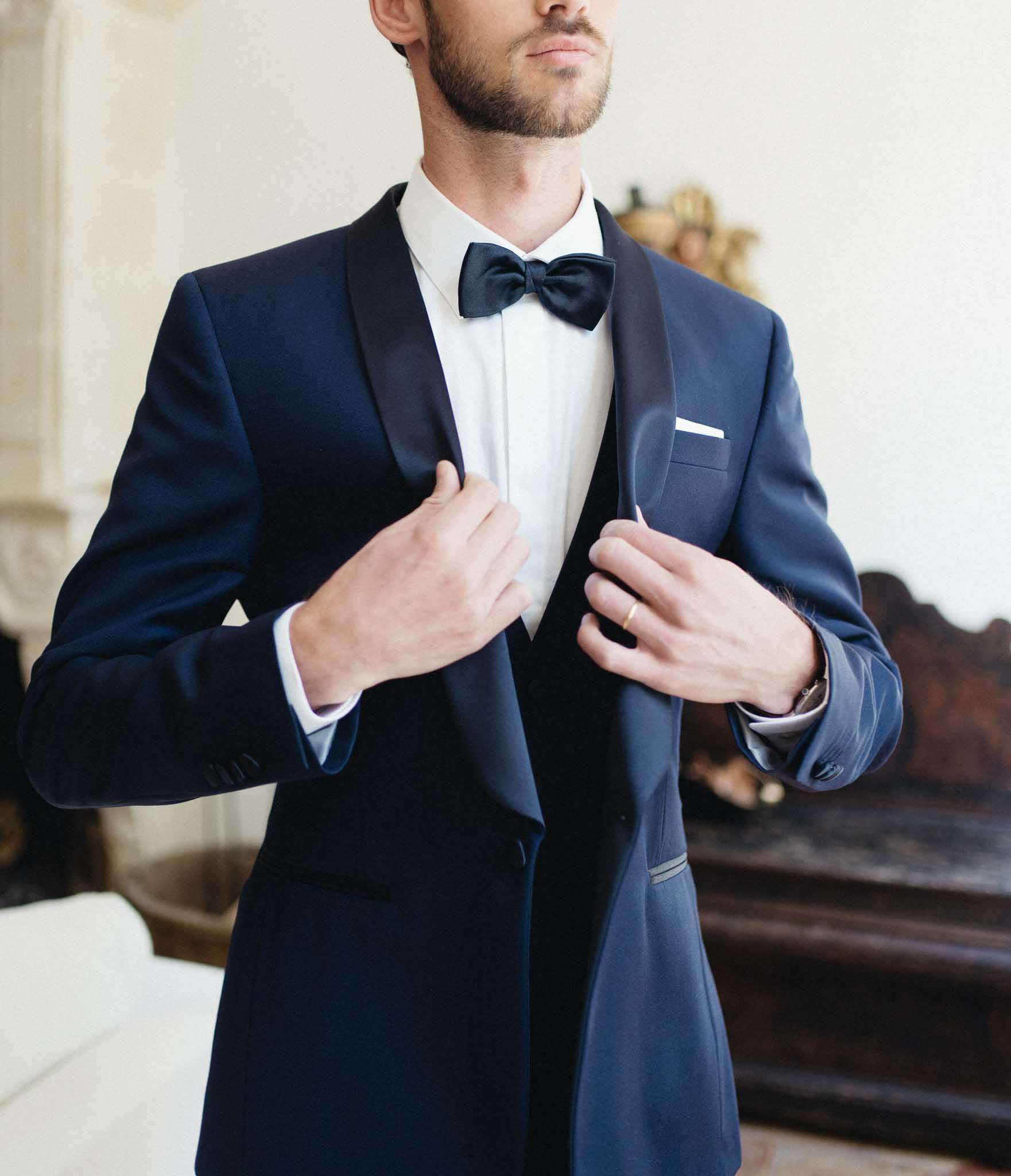 A getting-ready portrait of a groom adjusting the lapels of his navy blue tuxedo jacket, which features black satin shawl lapels and a matching white pocket square. He wears a white dress shirt, a navy satin bow tie, and a gold wedding band is visible on his right hand. The shot is a close-up torso portrait, cropped just below the chin and above the knees, with an ornate dark wood antique console or fireplace surround visible in the background, suggesting an interior room within a château or historic French property.