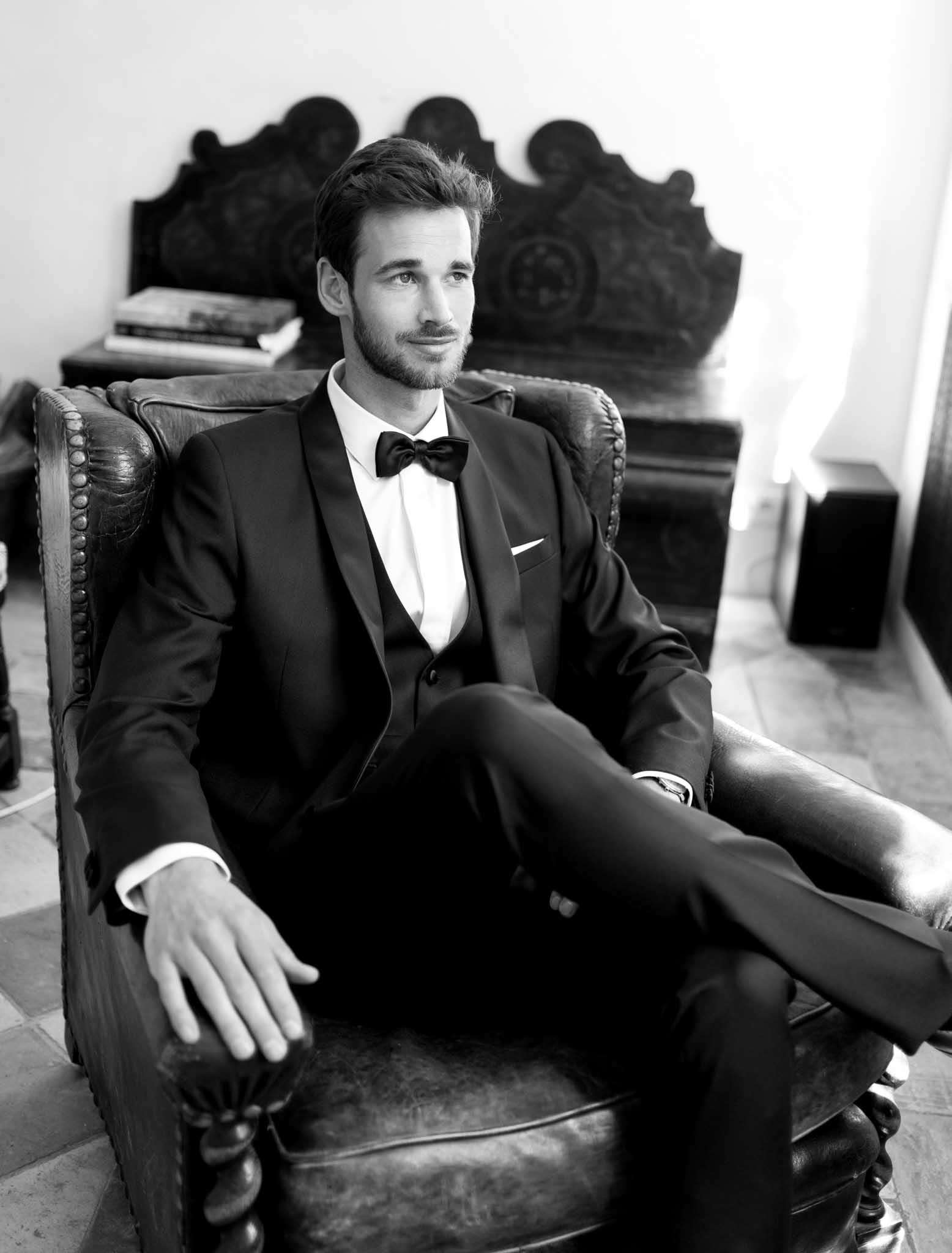 Black-and-white portrait of a groom seated in a worn leather wingback armchair indoors, shot in three-quarter length. He is wearing a black tuxedo with satin lapels, a white dress shirt, a black bow tie, and a white pocket square, and has a short beard. His legs are crossed and he looks slightly off-camera with a relaxed expression. Behind him is an ornately carved wooden bench or settle, and a small side table holding a stack of books. The tiled floor and plastered wall suggest a classic or rustic interior setting. The image has strong contrast with bright highlights from a nearby window. Portrait composition with shallow depth of field.