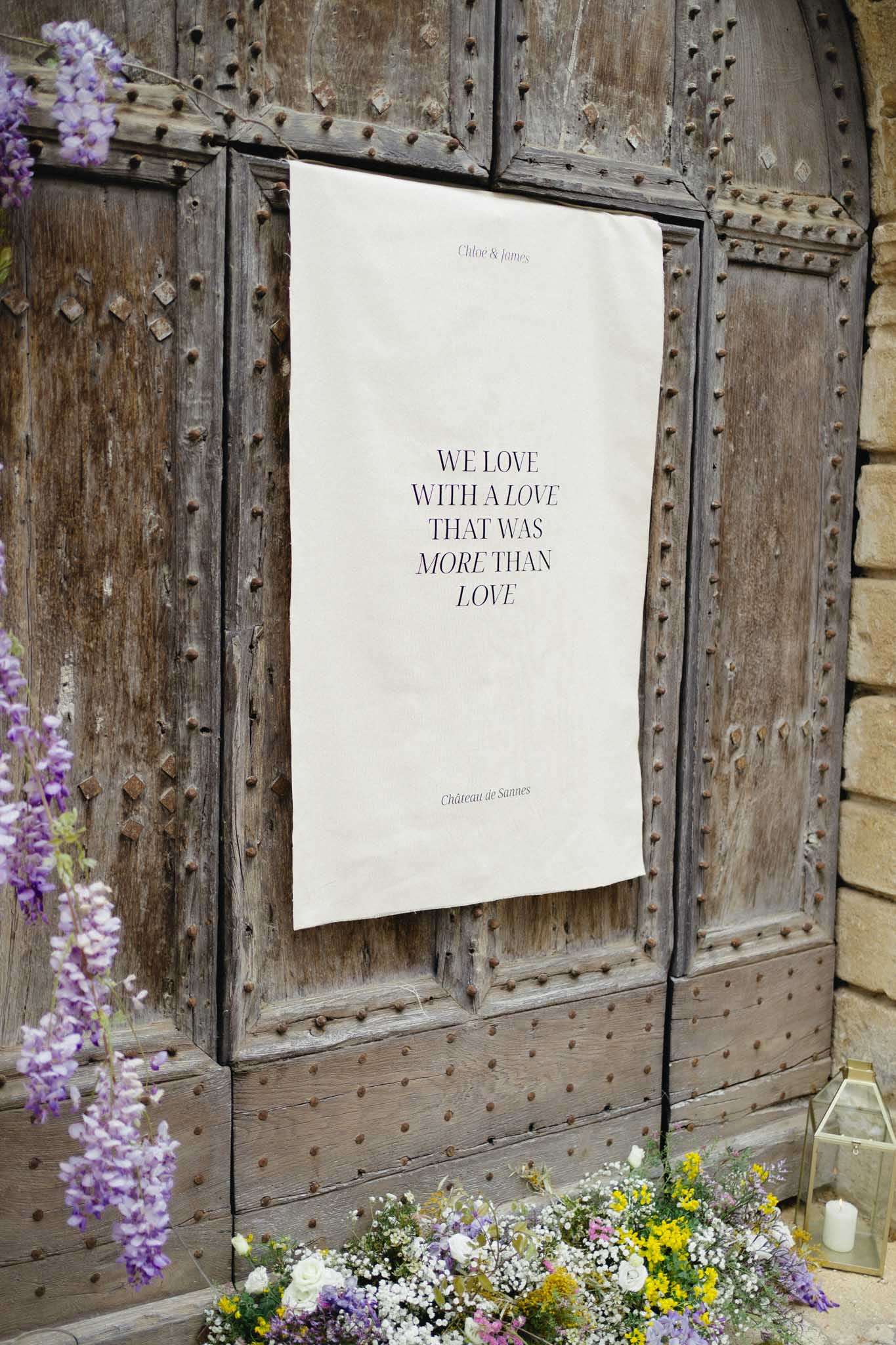 Wedding welcome banner on studded wooden door with roses, mimosa, lavender, and wisteria at Chateau de Sannes
