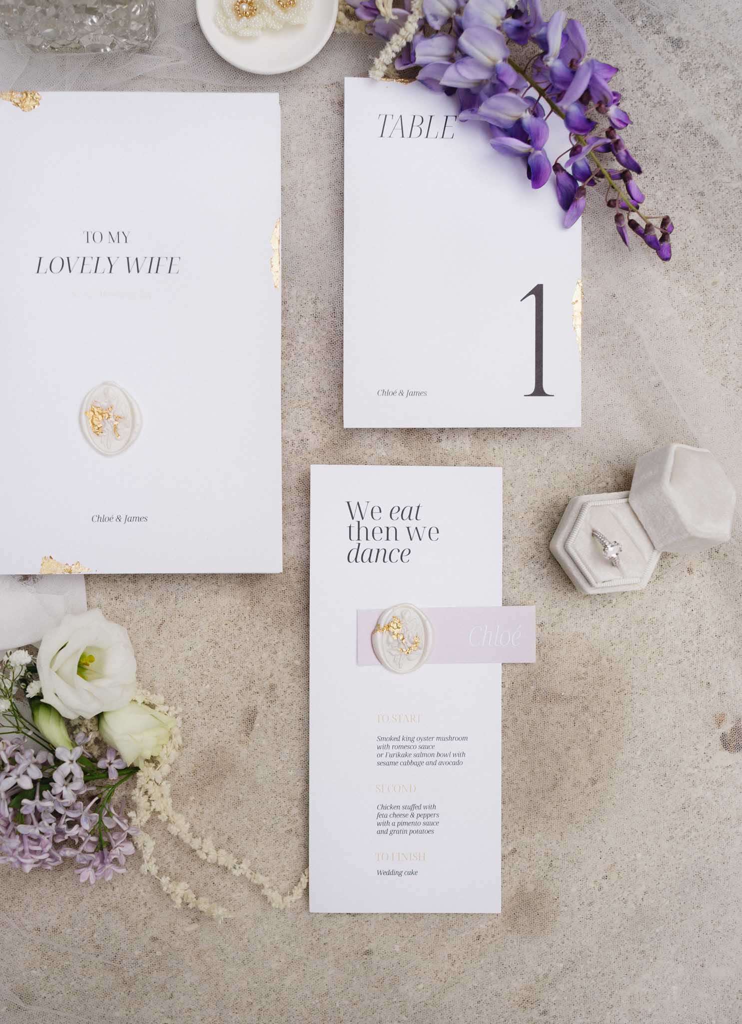 Stationery flat lay with gold foil vow booklet wax seals lavender menu card diamond ring and wisteria sprigs