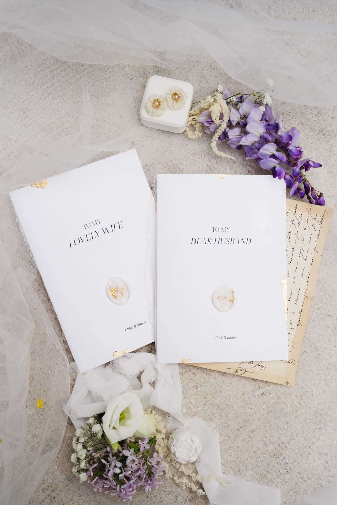 A flat lay detail shot of wedding stationery and accessories arranged on a neutral stone-toned surface. Two white vow booklets are displayed side by side — one reading 'To My Lovely Wife' and the other 'To My Dear Husband,' both personalised with the names Chloé & James and sealed with a white wax seal featuring gold floral detailing and gold corner accents. Surrounding the booklets are a white velvet jewelry box containing pearl and gold stud earrings, sprigs of purple wisteria and lilac blooms, small white lisianthus and astilbe flowers, aged handwritten letters, a sheer white veil, and white silk ribbon. The overall styling palette is white, gold, and soft purple, with a classic and romantic stationery aesthetic.