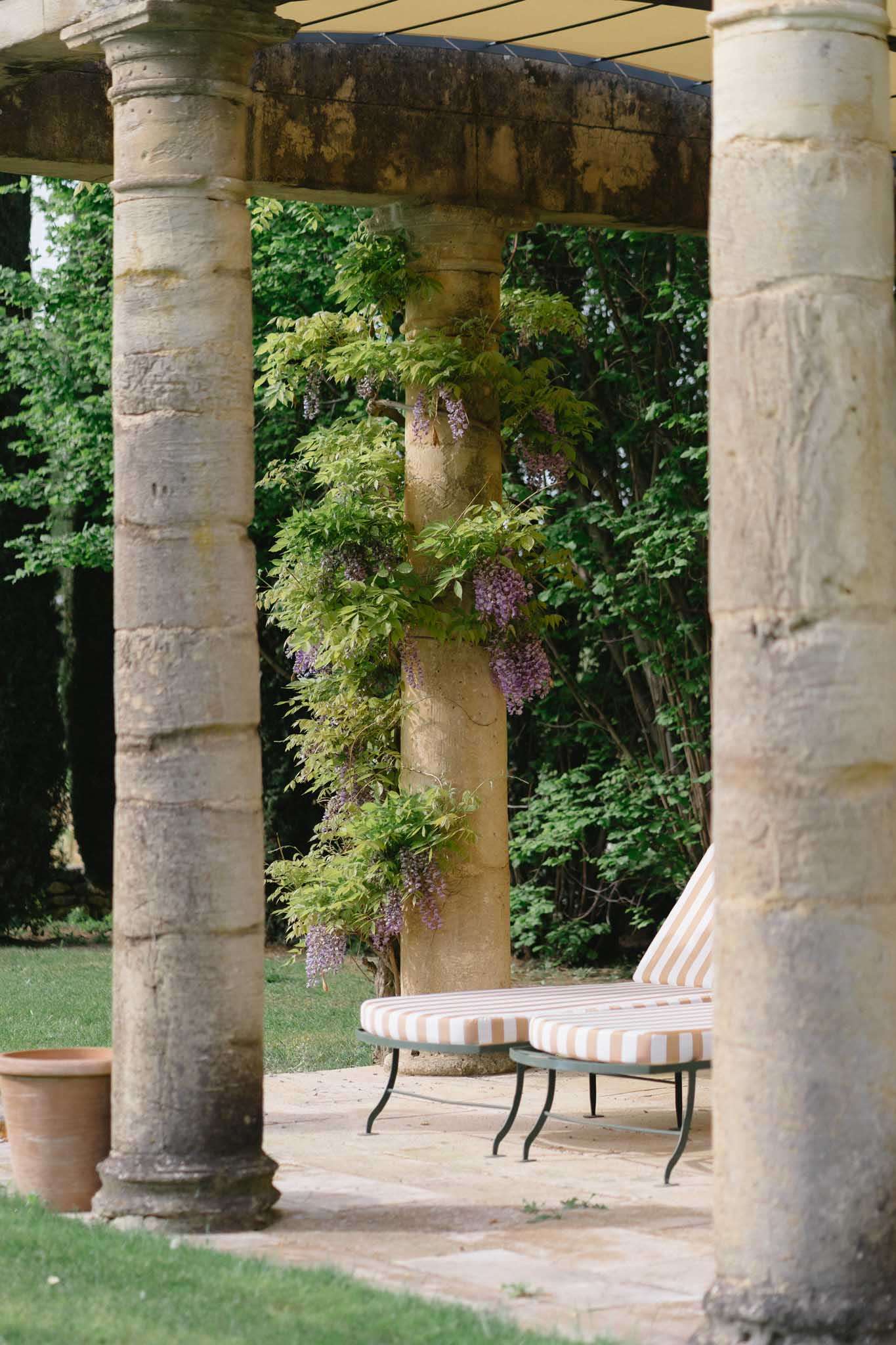 An outdoor covered terrace or loggia at what appears to be a French country estate or château, photographed as a venue detail shot. The image shows aged stone columns supporting an overhead structure, with purple wisteria climbing and trailing down one of the central columns. A chaise longue with a terracotta and ivory stripe cushion on a dark wrought iron frame is positioned between the columns on a stone-paved floor. A terracotta pot is visible to the left edge of the frame. The composition is a medium wide shot framed between two foreground columns, with a manicured lawn and garden visible in the background. No people are present. Potential venue feature image.