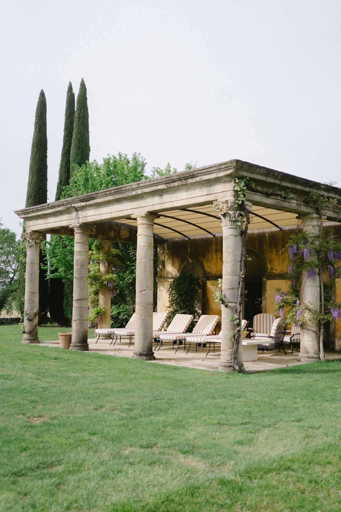 Stone colonnade pavilion with Corinthian columns and purple wisteria climbing beside sun loungers