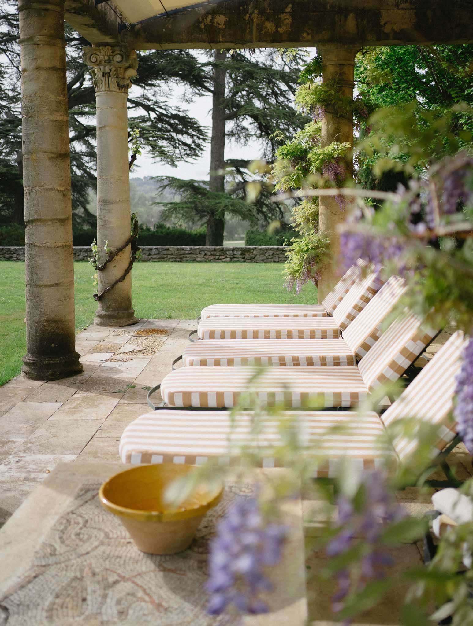 A wide shot of an outdoor terrace at what appears to be a French château or manor, featuring a row of sun loungers with tan and white striped cushions lined up under a covered colonnade with ornate stone Corinthian-style columns. Wisteria with purple blooms climbs the columns on the right side, and in the foreground a mustard yellow ceramic bowl sits on a mosaic stone floor detail. The terrace looks out over a manicured lawn with a low stone boundary wall and large cedar trees in the background. No people are visible; this is a venue detail shot captured with a shallow depth of field, with the wisteria in the foreground slightly out of focus. Potential venue feature image.