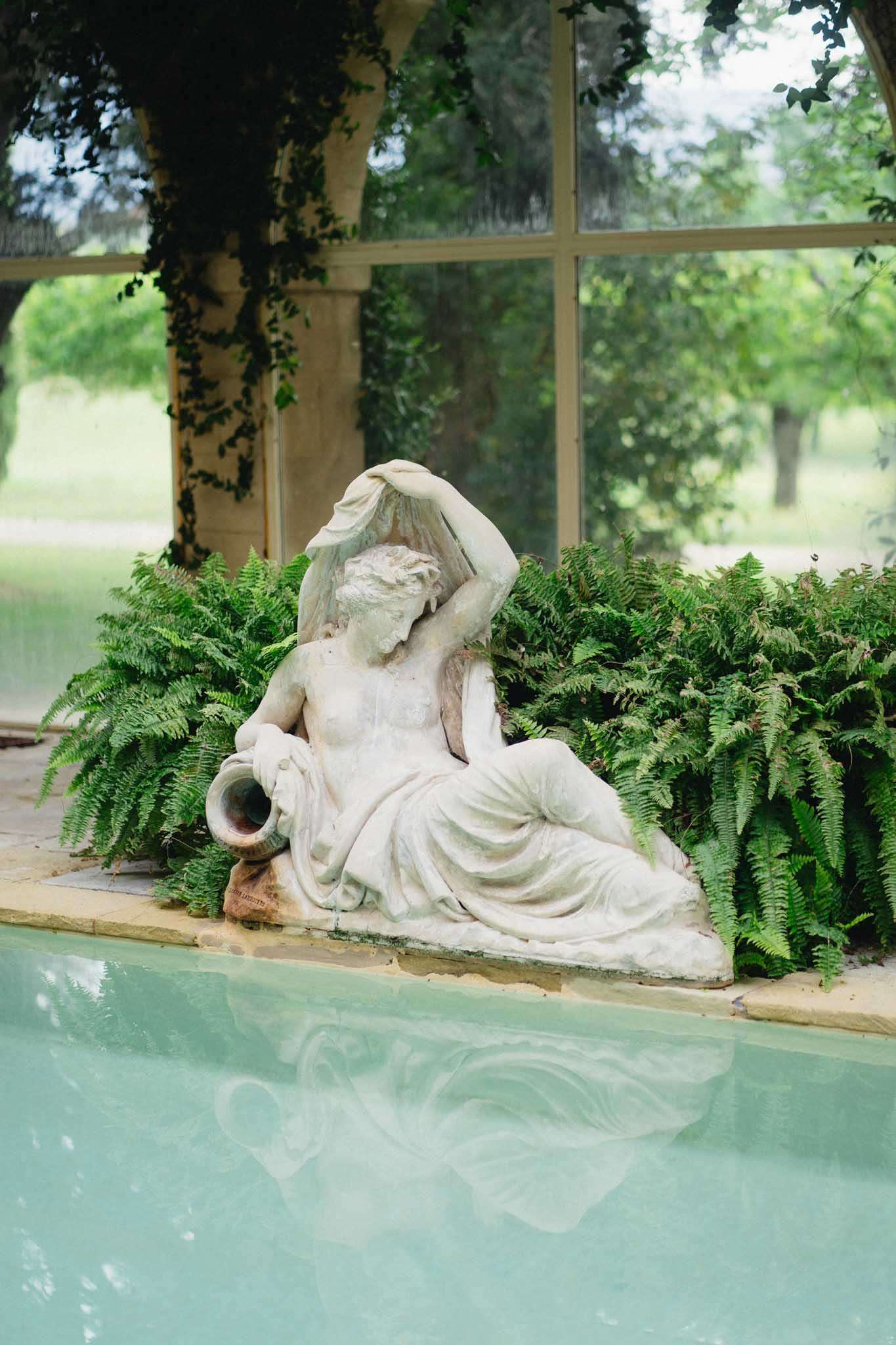 White stone sculpture of reclining female figure beside a turquoise reflecting pool with ferns and ivy-covered glass windows