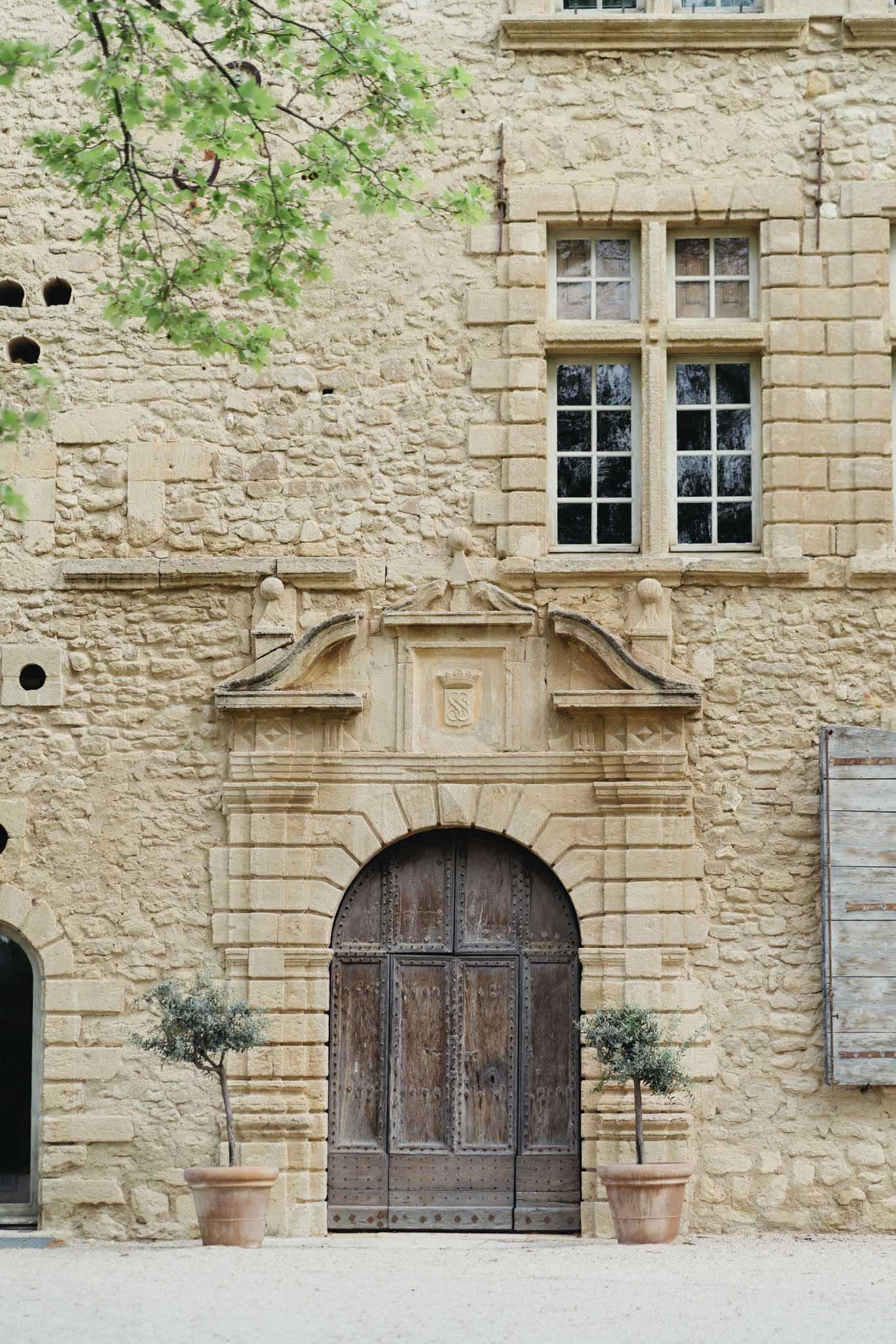Honey-toned limestone chateau facade with arched wooden double door, coat of arms pediment, and flanking topiary olive trees