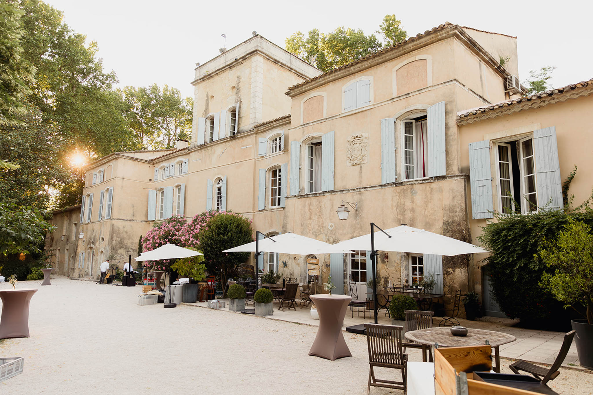 A wide shot of the exterior courtyard of a classic Provençal mas or château, set up for a wedding cocktail hour. The multi-story cream-rendered building features pale blue-grey shutters, terracotta roof tiles, and a carved stone coat of arms above the central entrance. The gravel courtyard in front of the building is furnished with cocktail-height tables covered in taupe stretch linen, white cantilever parasols, and wooden bistro-style chairs and round tables. Two staff members in white shirts are visible near a bar setup along the left wing of the building. Potted topiary boxwood shrubs and pink flowering oleander add to the outdoor décor. The warm golden light of late afternoon sun flares in from the left side of the frame. Potential venue feature image.