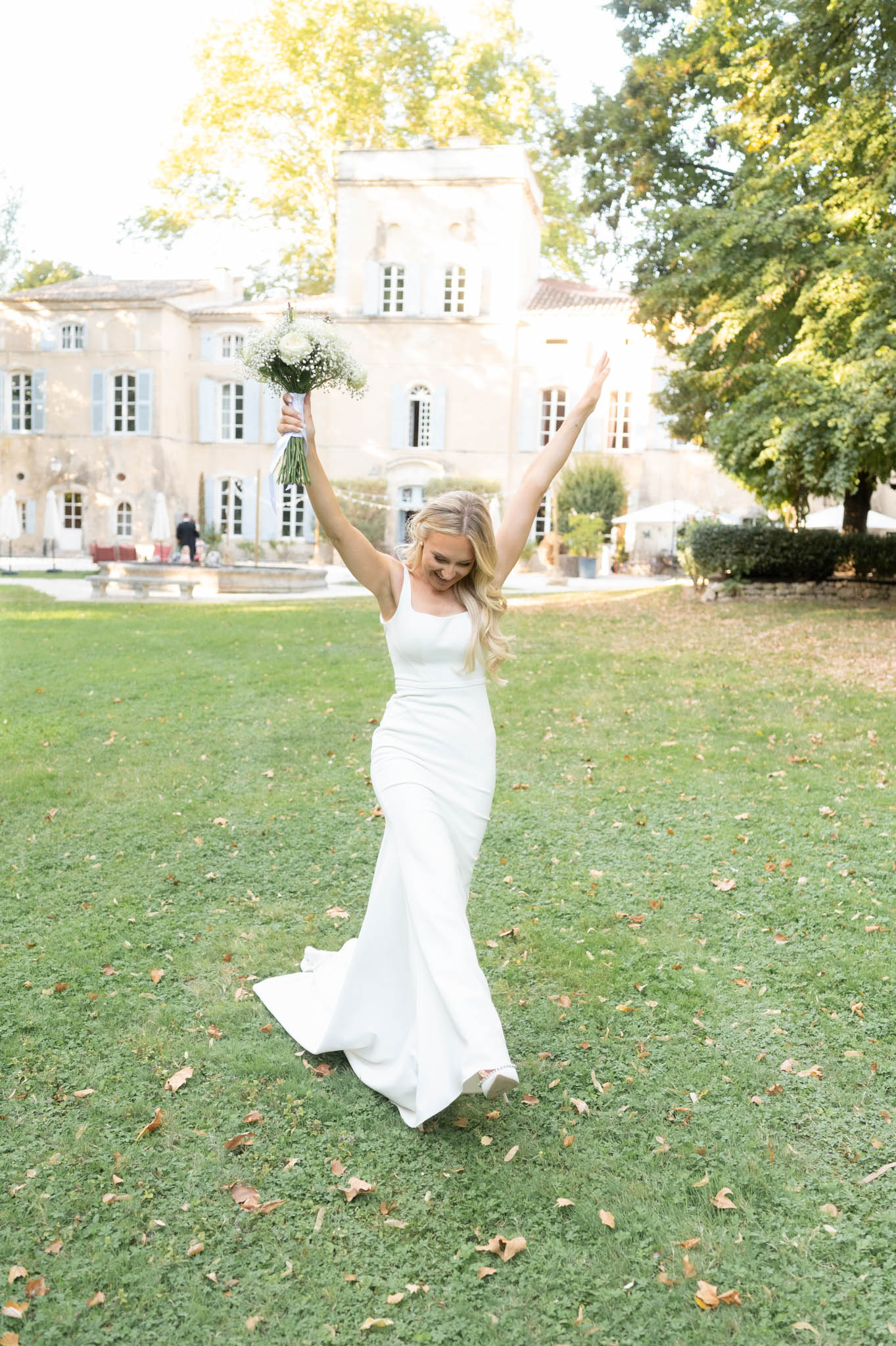 Bride laughing with bouquet raised on chateau lawn wearing ivory satin gown with Provencal manor behind