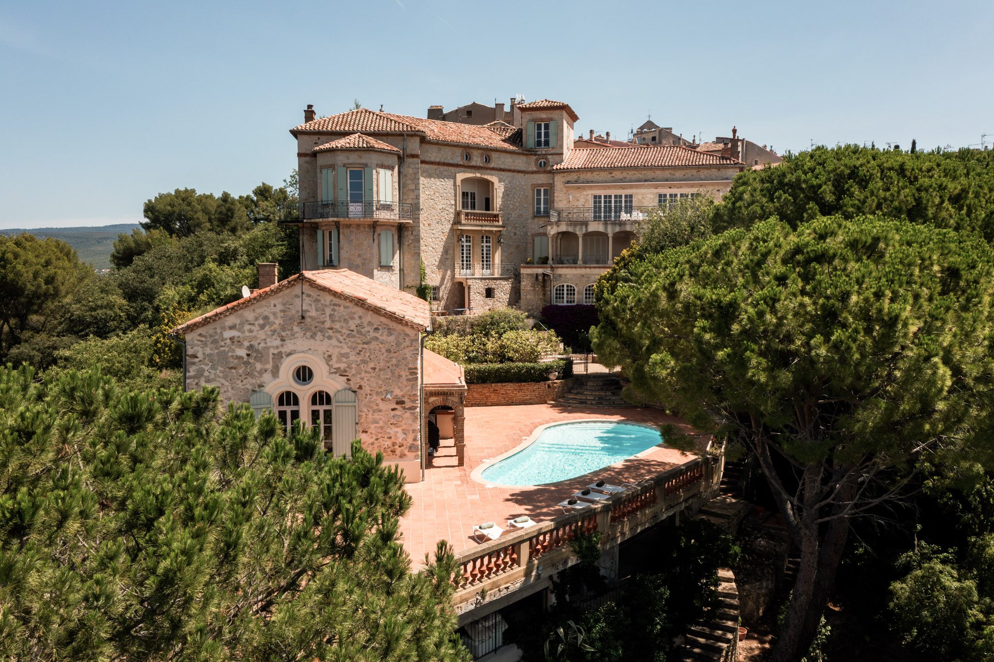 Aerial view of a Provencal stone manor with terracotta roof, pale blue shutters, pool, and pine tree grounds