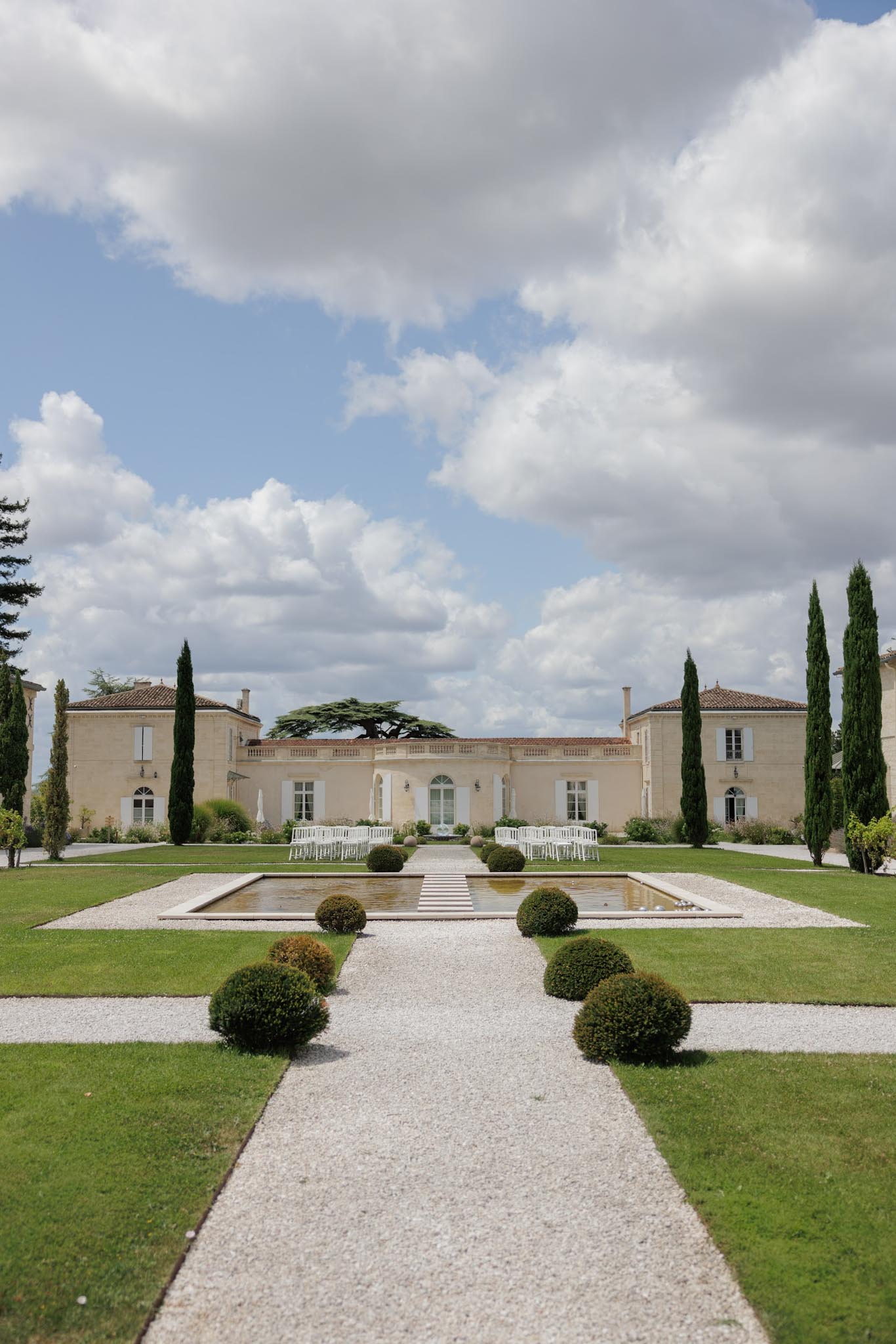 French chateau facade with formal garden, reflecting pool, trimmed hedges, and white ceremony chairs on gravel pathway