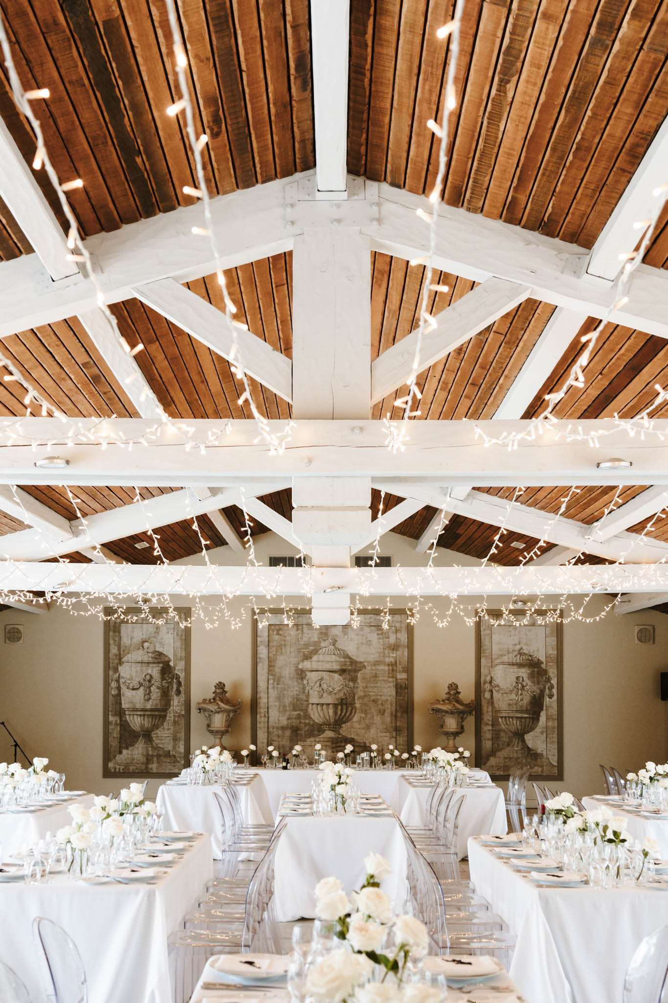 A wide shot of an indoor wedding reception room set up for a seated dinner. The venue features an exposed vaulted ceiling with warm honey-toned wooden planks and white-painted structural beams, draped with warm white fairy lights that cascade downward. Three long rectangular dining tables are dressed in white linen tablecloths and set with glassware, white plates, and white napkins. Clear acrylic ghost chairs line both sides of each table. Centerpieces consist of low arrangements of white roses and white blooms in glass vessels, with single-stem white roses in bud vases spaced along the tables. The back wall displays three large antique-style sepia-toned painted panels depicting classical stone urns, contributing to a classic, refined decor palette of white and neutral tones. The overall styling is classic and understated with a white-on-white floral and linen scheme. Potential venue feature image.