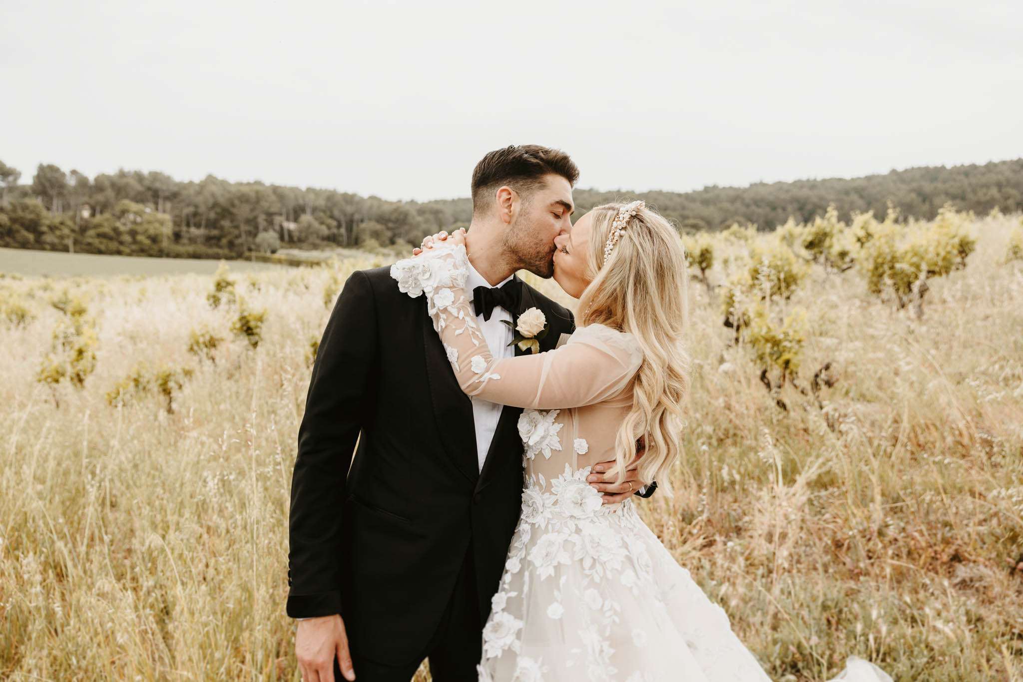 Bride in floral applique tulle gown kissing groom in black tuxedo by vineyard