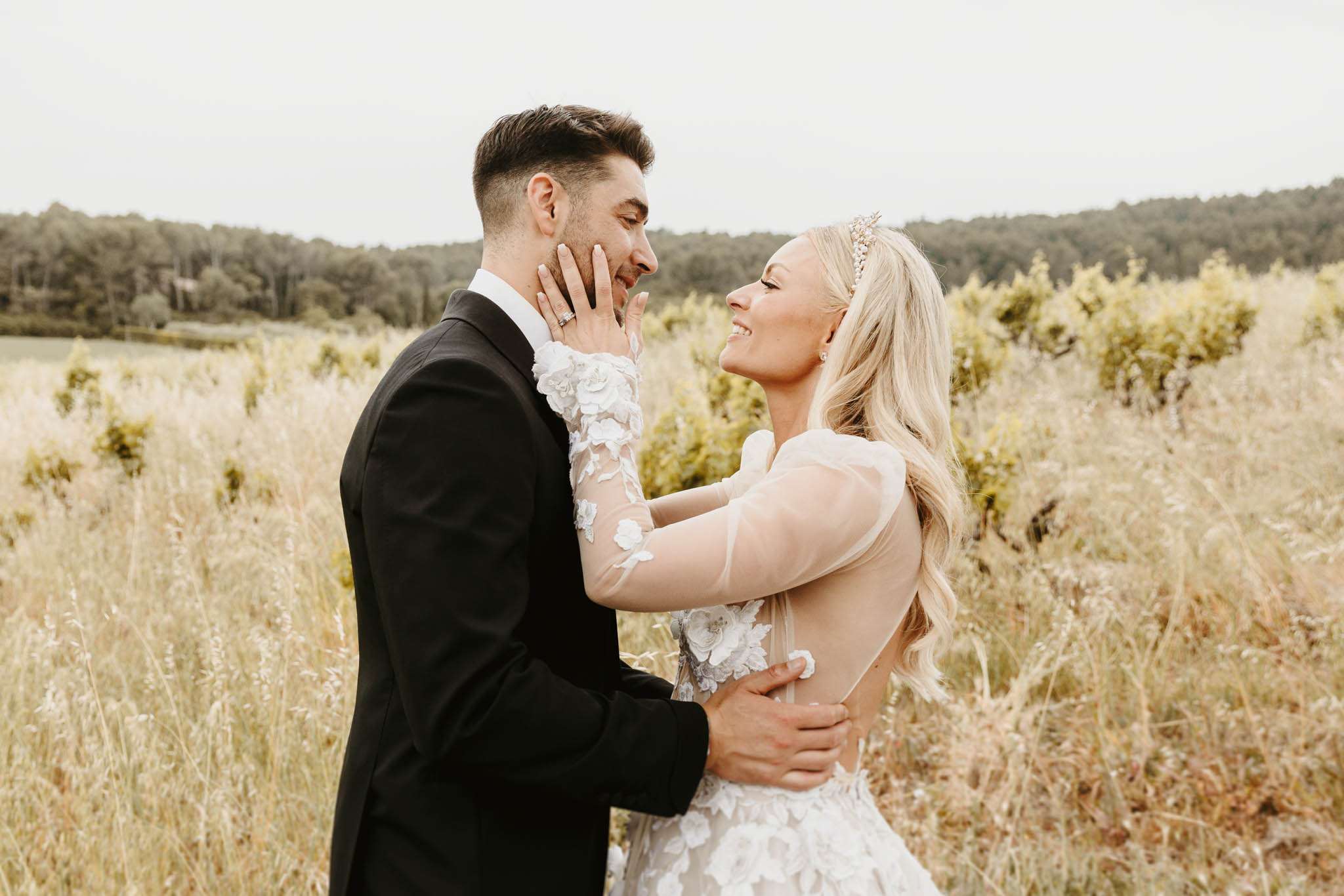 Bride in sheer long-sleeve gown with floral appliques and crystal headband embracing groom in black tuxedo