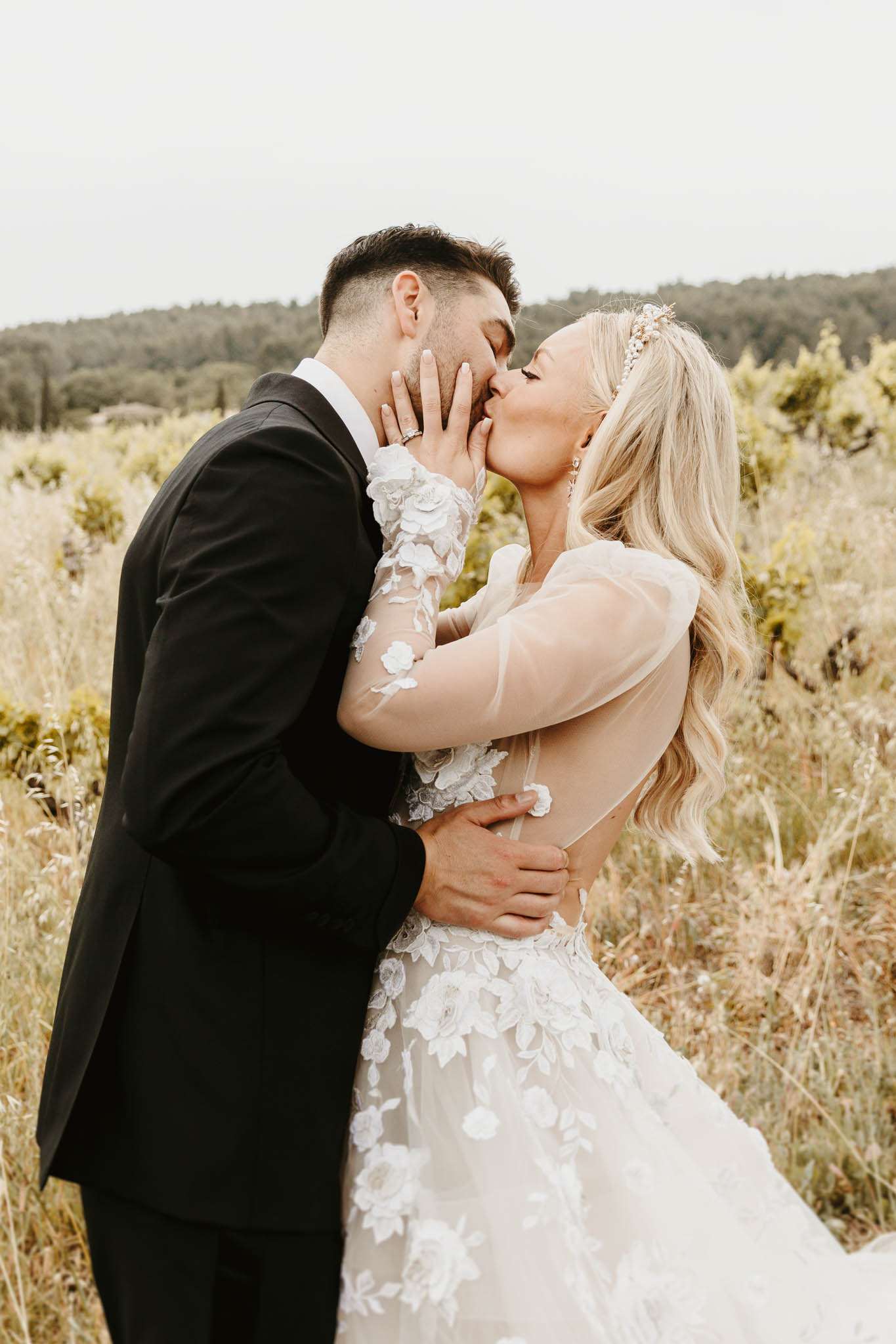 A couple portrait shoot outdoors, with the bride and groom kissing in a natural scrubland setting. The groom wears a black tuxedo jacket with a white dress shirt, while the bride wears a long-sleeve illusion tulle gown featuring white 3D floral appliqués across the skirt and sleeves, with a low open back. The bride has long blonde wavy hair accessorized with a pearl and crystal headband, and she places her hand gently on the groom's face during the kiss. The shot is a medium close-up portrait framing both figures from roughly the waist up, with a soft-focus natural background.