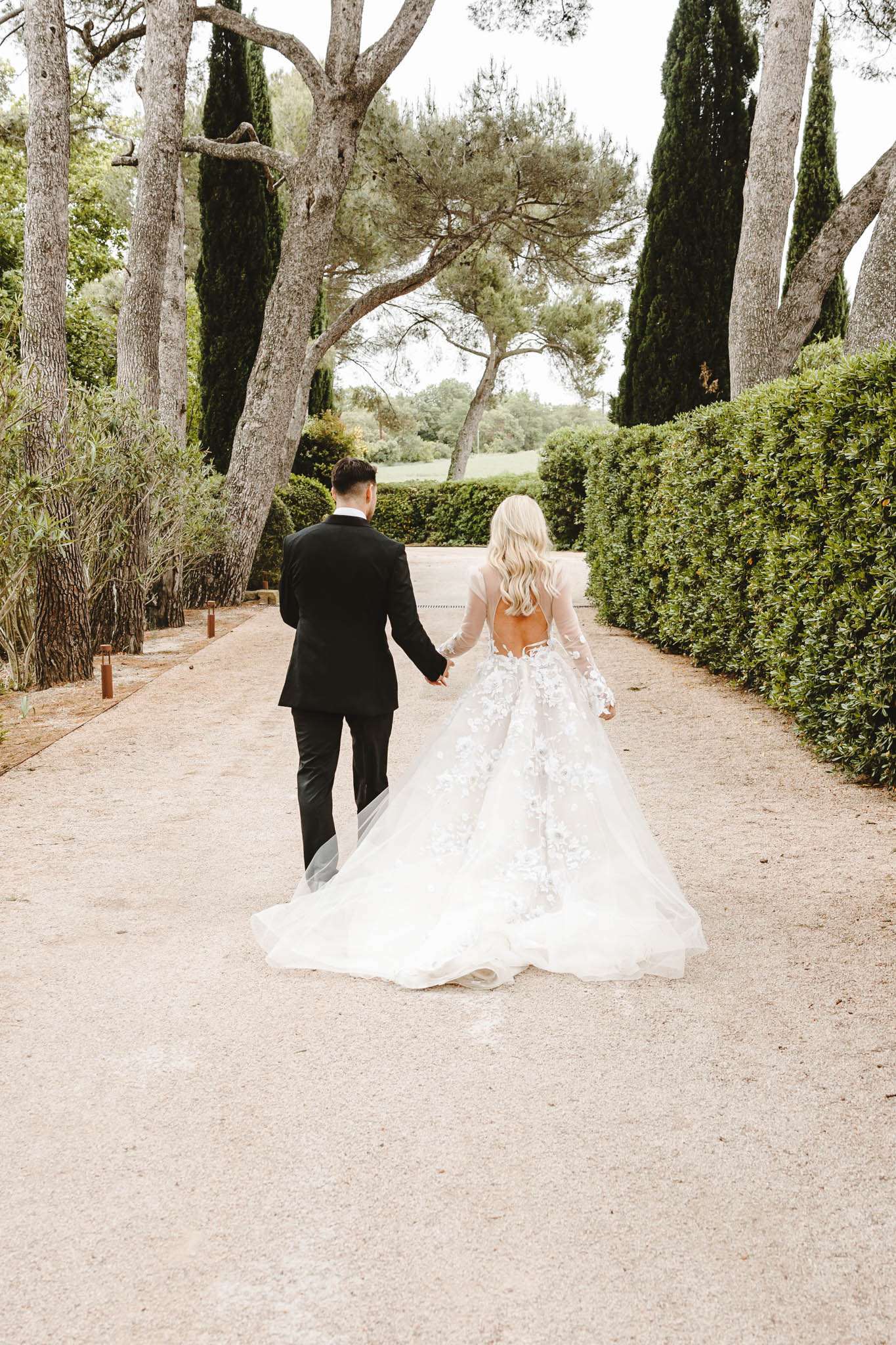 Bride and groom walking hand-in-hand along hedge-lined gravel path viewed from behind in formal garden