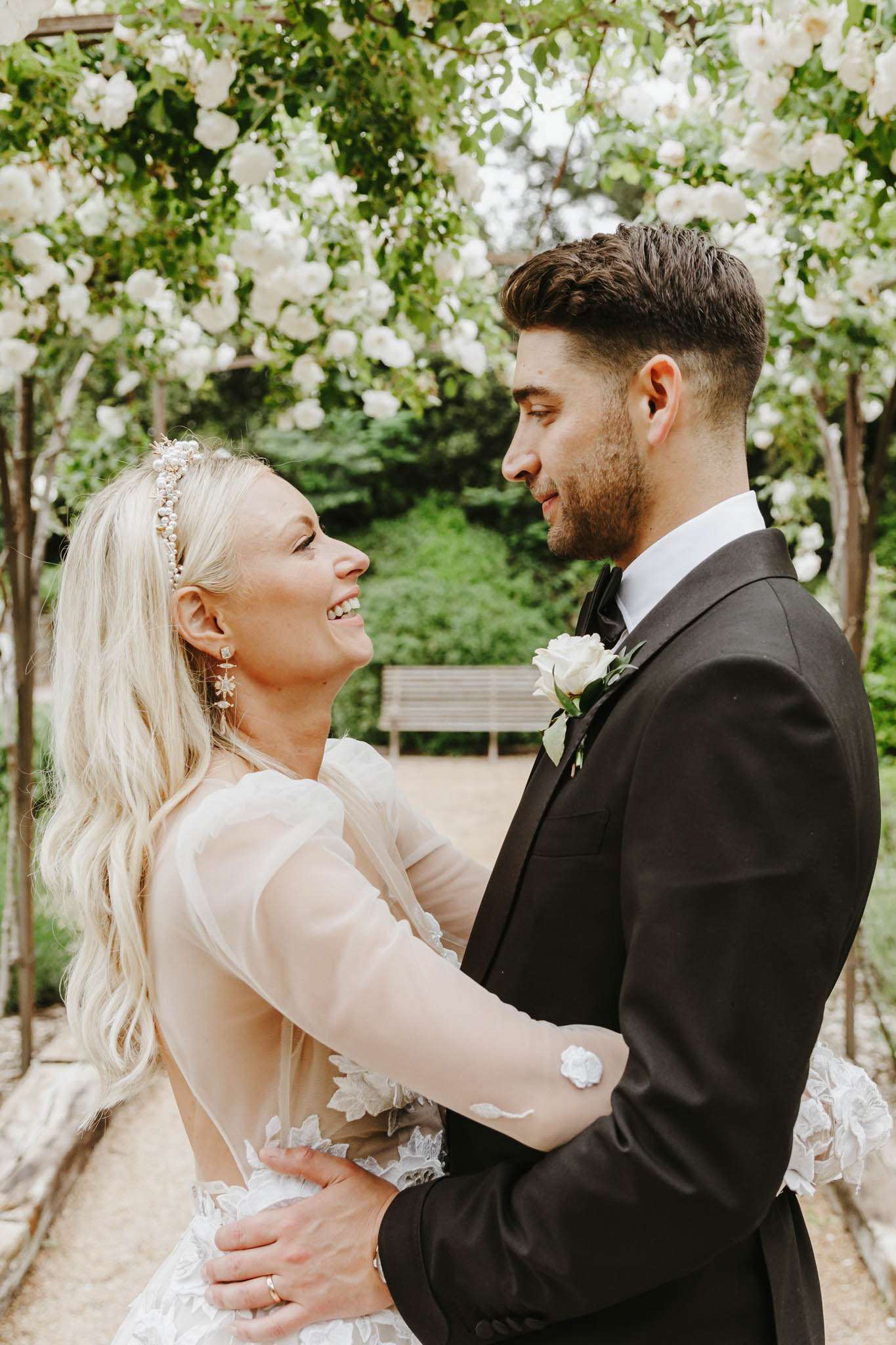 A couple portrait taken outdoors in a garden setting beneath a pergola or arch covered in white climbing roses. The bride wears a long-sleeve sheer tulle and lace gown with floral appliqué detailing and an open back, accessorized with a pearl and crystal headband and dangling floral earrings, with her blonde hair worn loose. The groom wears a black tuxedo with a black bow tie and a white rose and greenery boutonniere. The two are facing each other with their arms around one another, the bride smiling broadly. A wooden garden bench is visible in the background along a gravel pathway. The overall styling is classic and garden-inspired, with a white and green color palette. Portrait crop, mid-shot framing.
