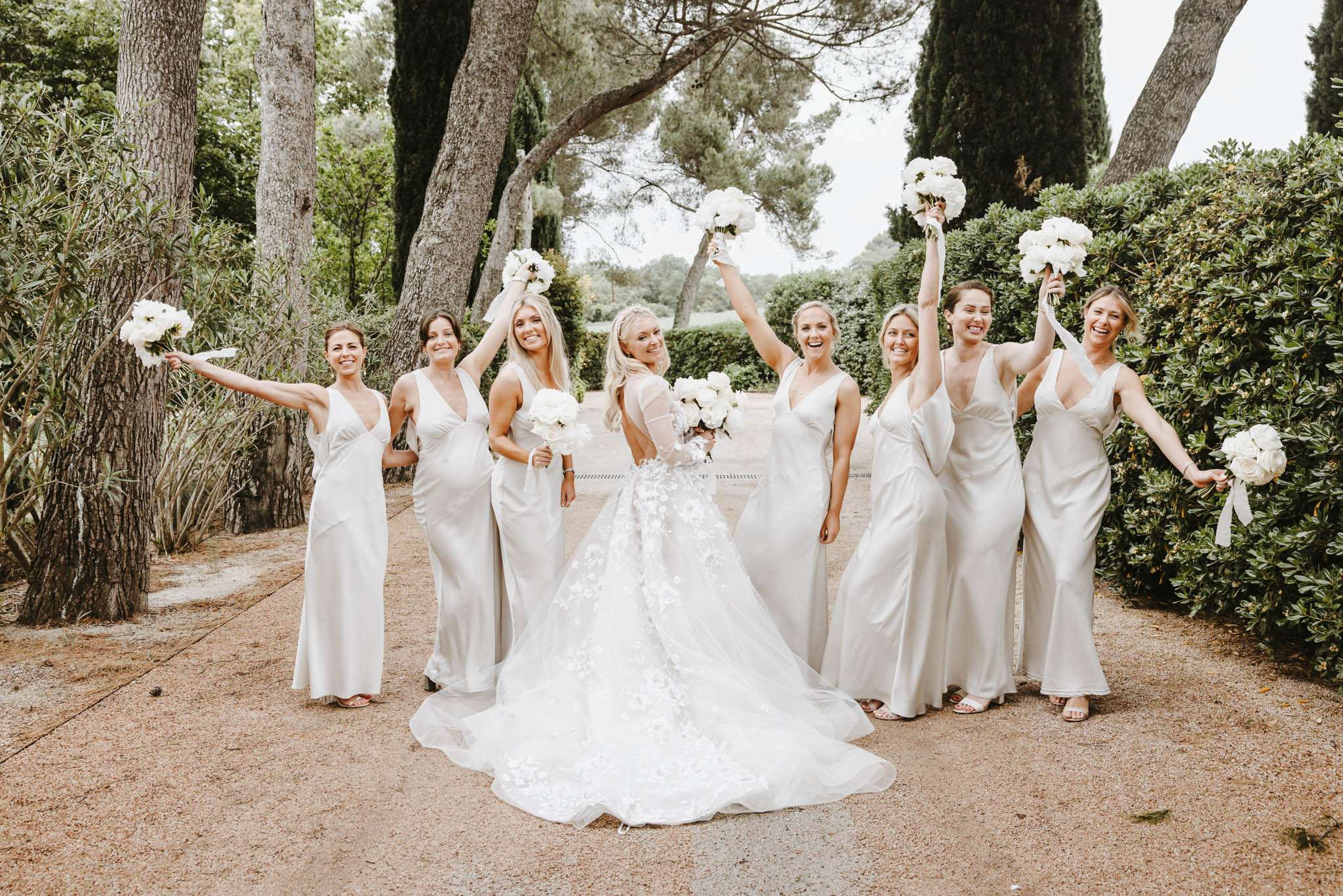 Bride in white floral ballgown from behind with seven bridesmaids in champagne satin gowns raising bouquets