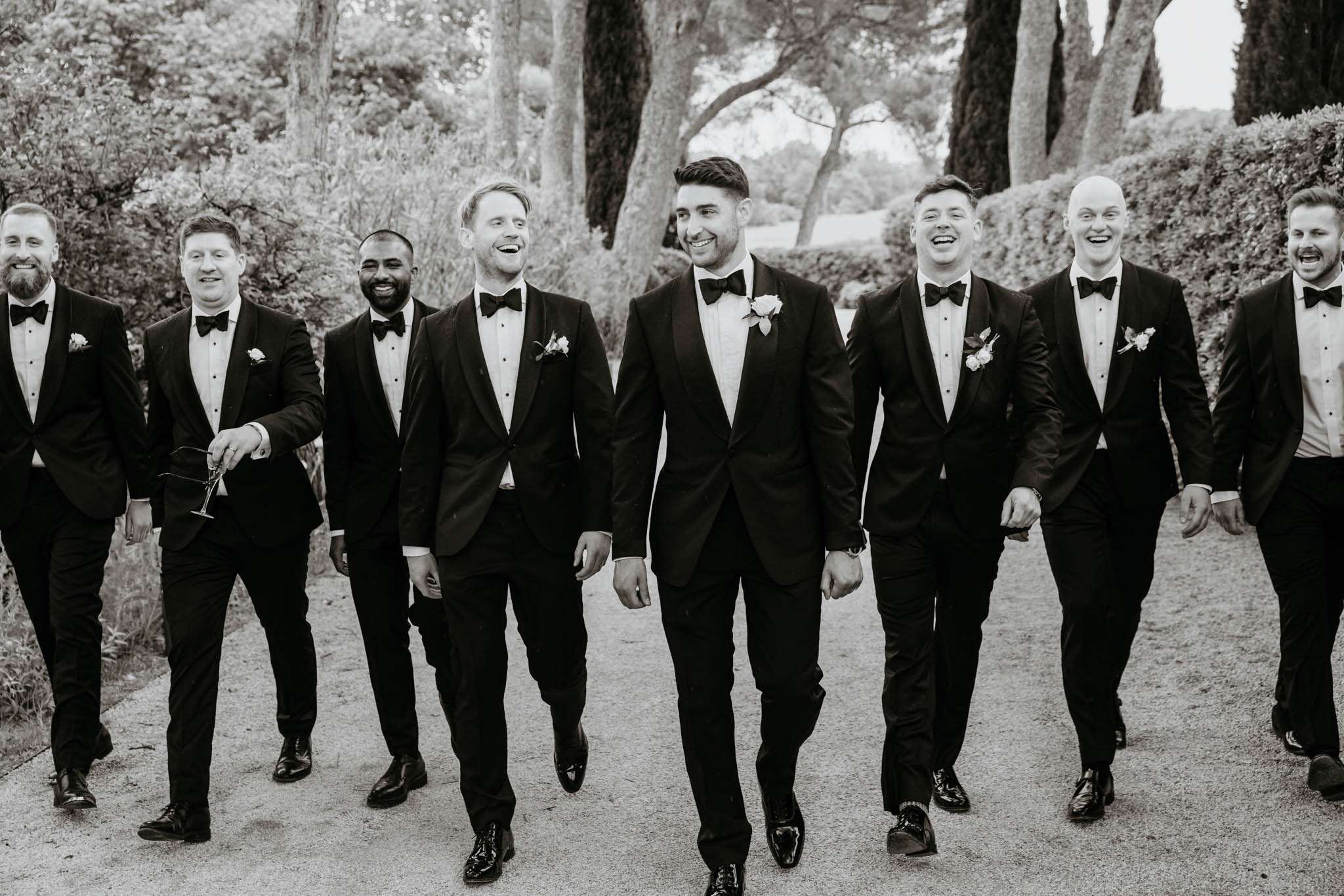 Black and white portrait of groom and seven groomsmen in tuxedos walking on tree-lined path