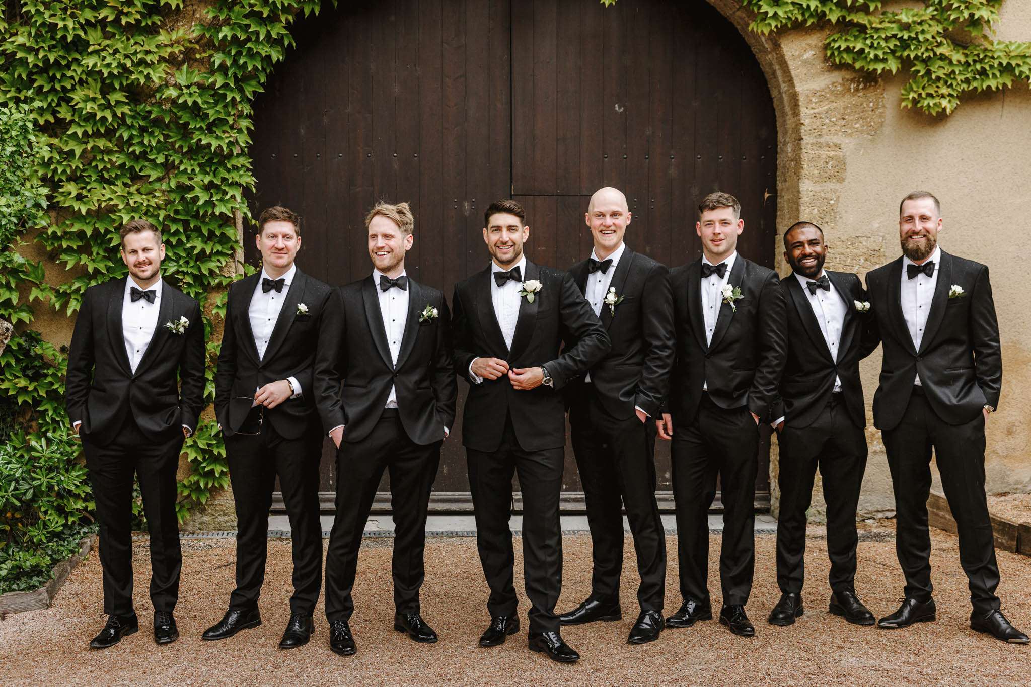 A group portrait of a groom and seven groomsmen standing in a row outdoors in front of a large arched wooden door set into a stone wall. All eight men are dressed in black tuxedos with white dress shirts and black bow ties, each wearing a white floral boutonniere — appearing to be small white roses or spray roses with minimal greenery. The groom stands at center and is distinguished by his position and a slightly different tuxedo lapel style. The setting appears to be a French château or manor, with the stone archway and gravel courtyard visible. The shot is a wide group portrait taken at eye level with the full group in frame.