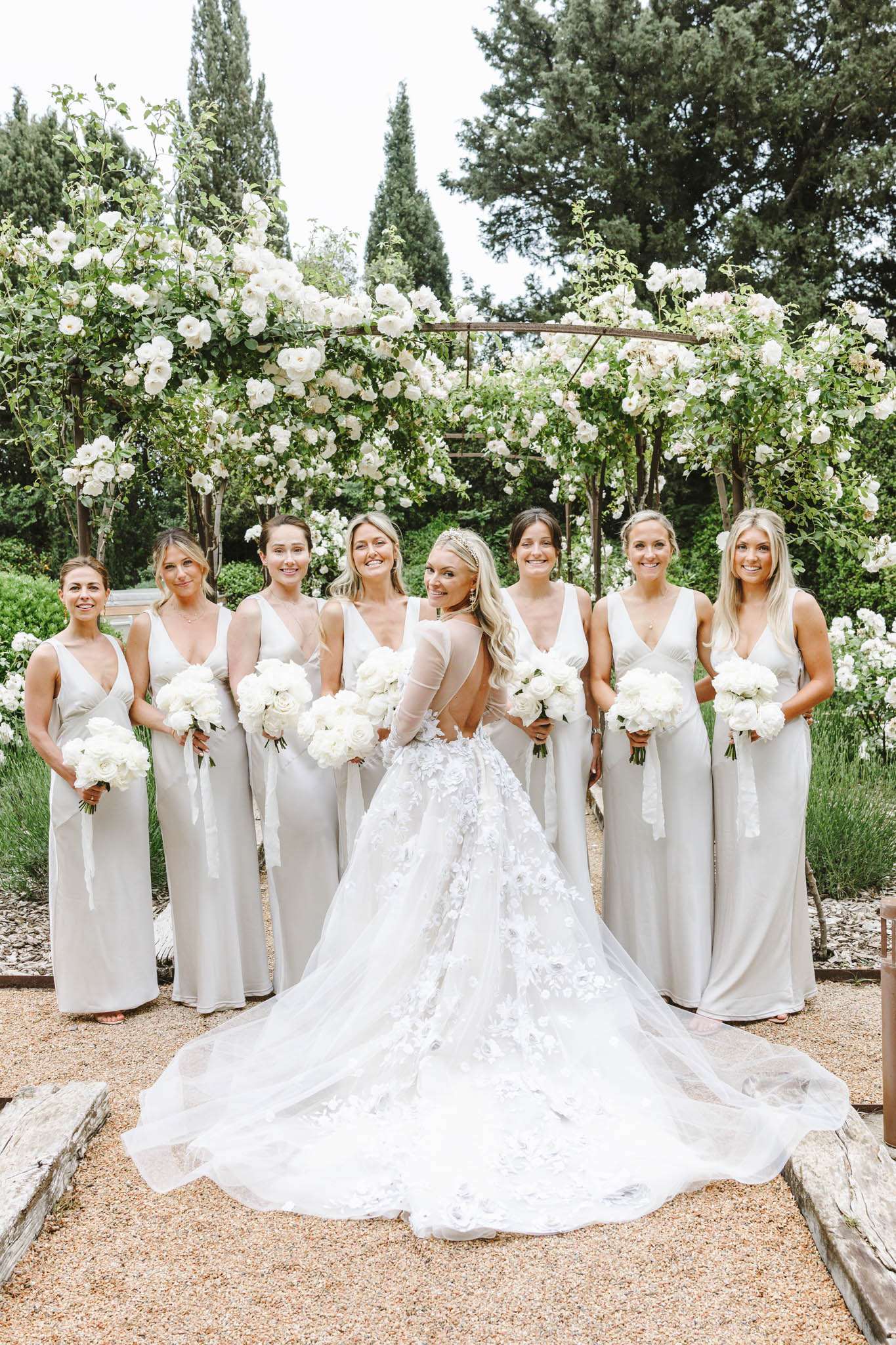 Bride and six bridesmaids in sage dresses with white rose bouquets before wooden rose pergola
