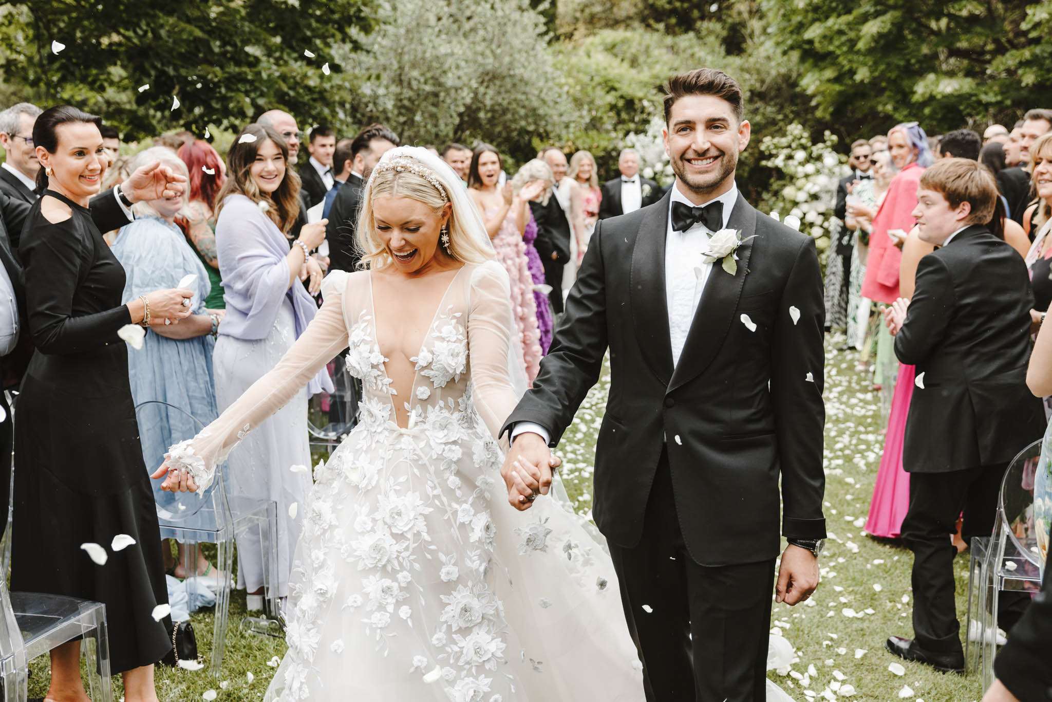 Bride and groom walk up the aisle as guests toss white petals after outdoor garden ceremony
