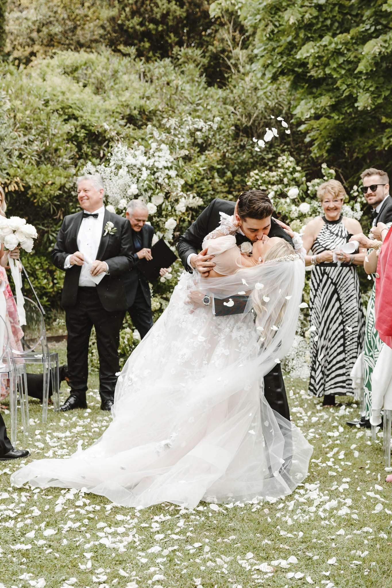 Dip kiss with petal toss under white rose arch with ghost chairs and scattered petals on grass