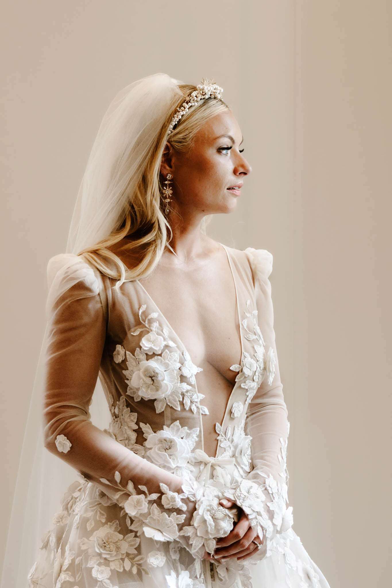 A close-up portrait of a bride shot against a plain warm beige background. She wears a deep V-neck wedding gown with a nude/blush illusion tulle base and long sleeves, covered in three-dimensional white floral and leaf appliqués. Her accessories include a gold and pearl floral headband crown from which a long ivory veil falls, and dangling gold celestial-style earrings. Her blonde hair is worn loosely down, and her hands are clasped in front of her. The composition is a three-quarter portrait from roughly waist height upward, with the bride's gaze directed off to the left.