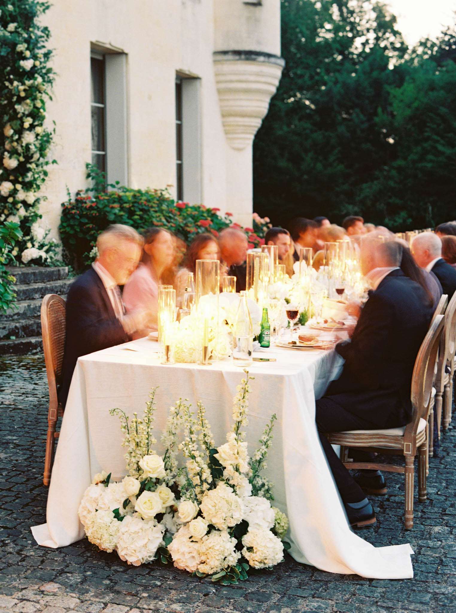 An outdoor wedding reception dinner is underway at dusk, with approximately 15–20 guests seated along a long rectangular table set on a cobblestone courtyard in front of a French château-style building. The table is dressed in a white linen tablecloth that pools slightly on the ground, and the centerpiece features a row of tall glass hurricane candle holders with warm candlelight running the length of the table, alongside white floral arrangements and glassware. At the base of the table, a lush floor-level floral arrangement spills forward, composed of white hydrangeas, ivory garden roses, white delphiniums, and greenery. Guests are dressed in formal attire — dark suits and evening wear — and the motion blur on several figures suggests a slower shutter speed was used; the wide shot is taken from a low angle that emphasizes the table length and foreground florals. Potential venue feature image.