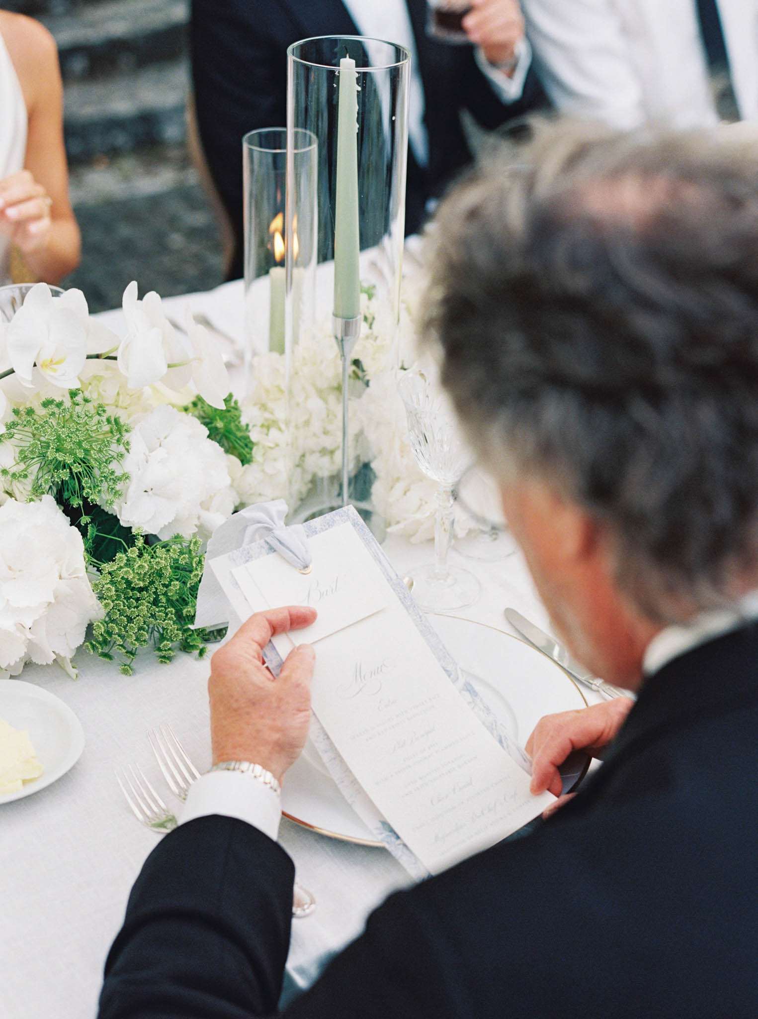 A close-up detail shot taken from above and behind a male guest seated at a wedding reception table, reading a calligraphed menu card. The place setting includes a gold-rimmed charger plate, silver cutlery, and crystal glassware on a white linen tablecloth. Centerpiece florals feature white hydrangeas, white orchids, and green ammi or bupleurum, arranged low and lush. Two tall glass hurricane vases hold sage green taper candles, one of which is lit. The menu card is printed with a light blue or lavender border and tied with a narrow ribbon, accompanied by a separate place card. The overall decor palette is white, green, and soft blue-grey with gold accents, reflecting a classic, refined reception style.