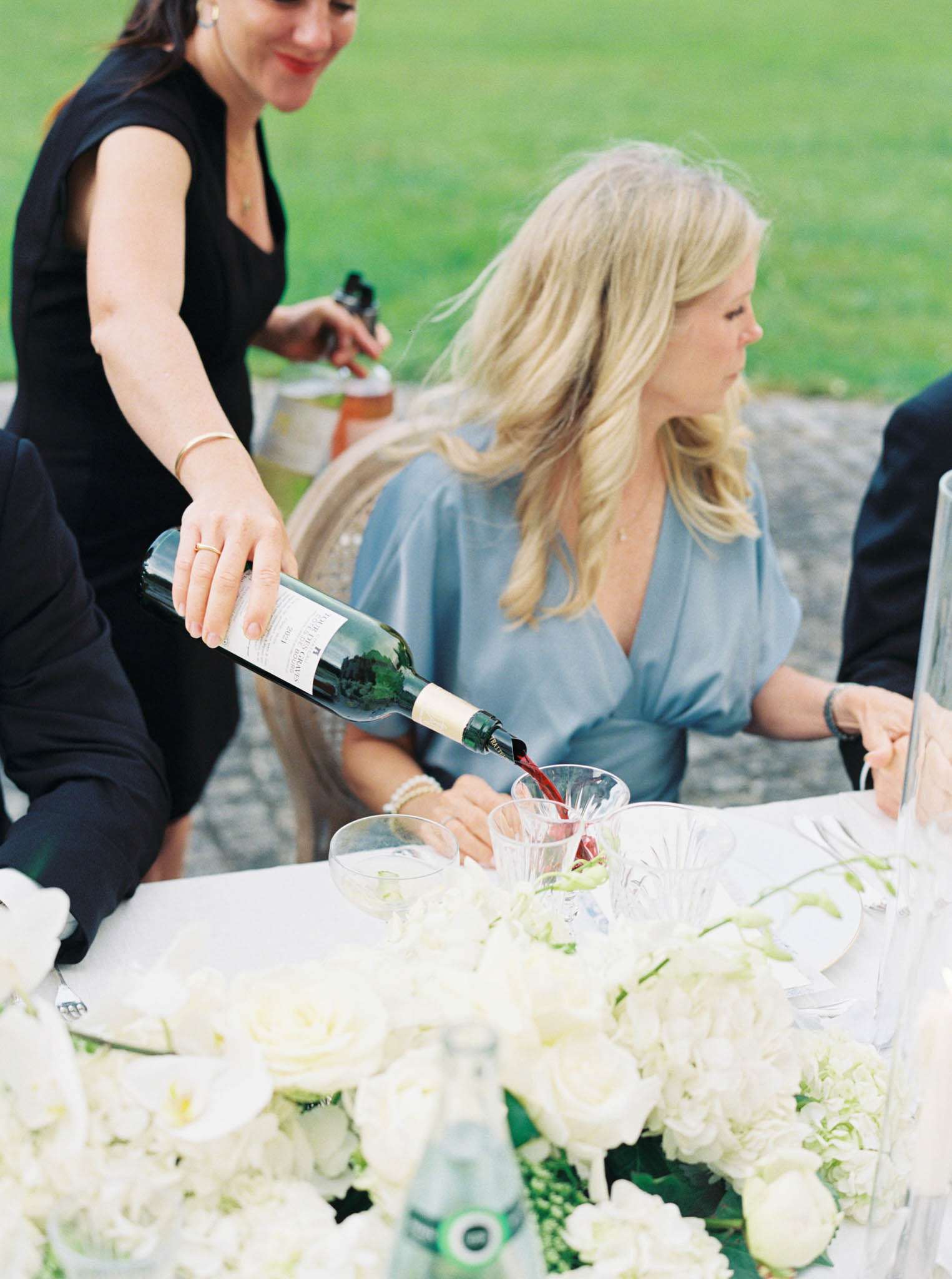 A candid close-up shot taken during an outdoor wedding reception, showing a server in black pouring red wine from a dark glass bottle into a crystal glass at the dining table. A female guest seated at the table is wearing a pale blue wrap-style dress and a pearl bracelet, with her hair loose. The white linen table is decorated with a lush low floral arrangement of white roses, white hydrangeas, and green accents, with crystal glassware and a small green glass water bottle visible among the blooms. At least two other guests are partially visible at the table, set against a green lawn in the background.
