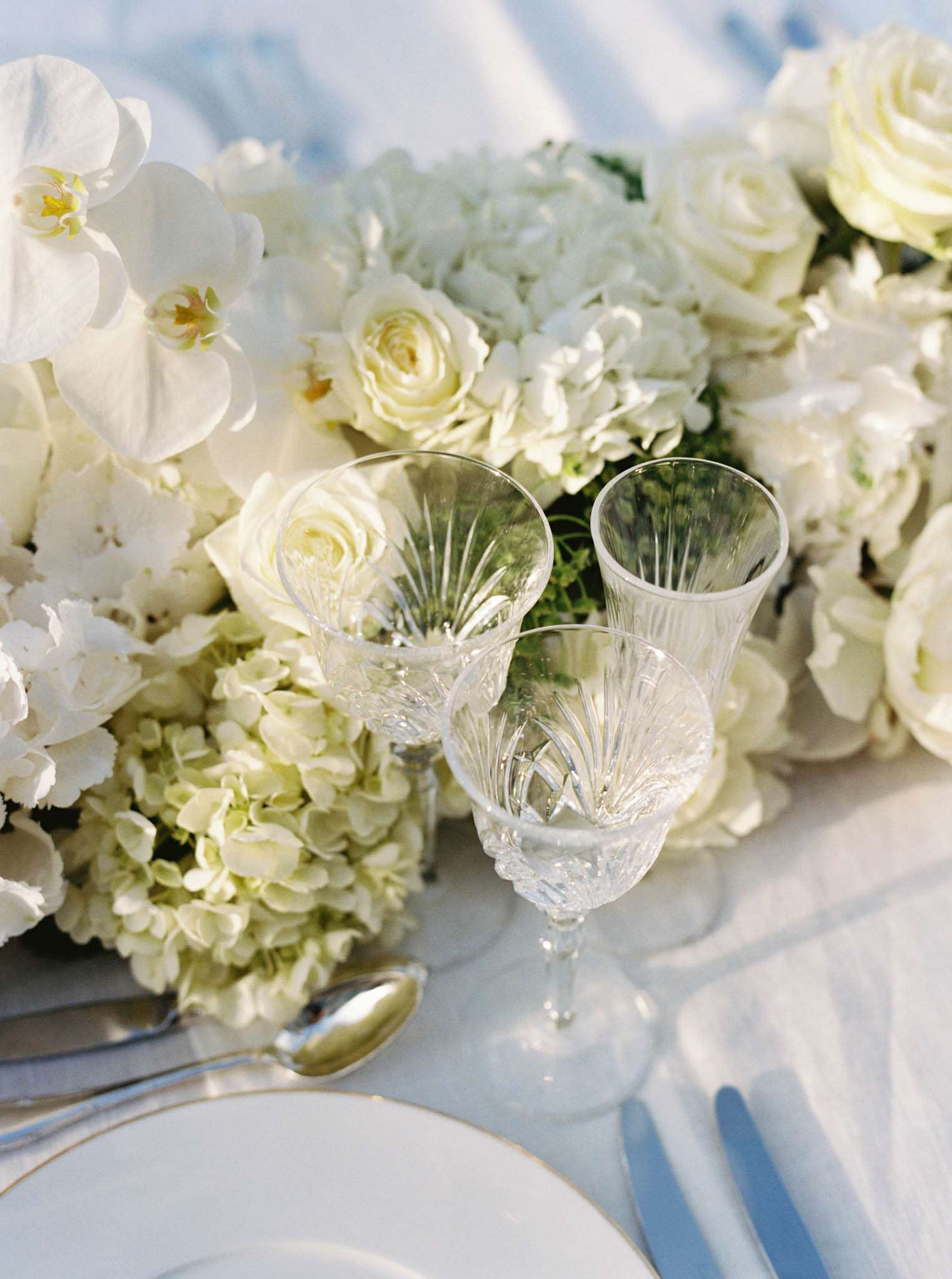 Close-up detail shot of a wedding reception tablescape featuring a lush all-white floral centerpiece composed of phalaenopsis orchids, garden roses, hydrangeas, and lisianthus arranged in a low, dense formation. Two cut-crystal goblets with ribbed detailing are positioned in front of the arrangement, partially tipped toward each other. The place setting includes a white plate with a thin gold rim, silver flatware including a visible spoon, and a pale blue-handled knife, all set on a white linen tablecloth. The overall decor palette is white, ivory, and soft blue, with a classic, formal styling approach.