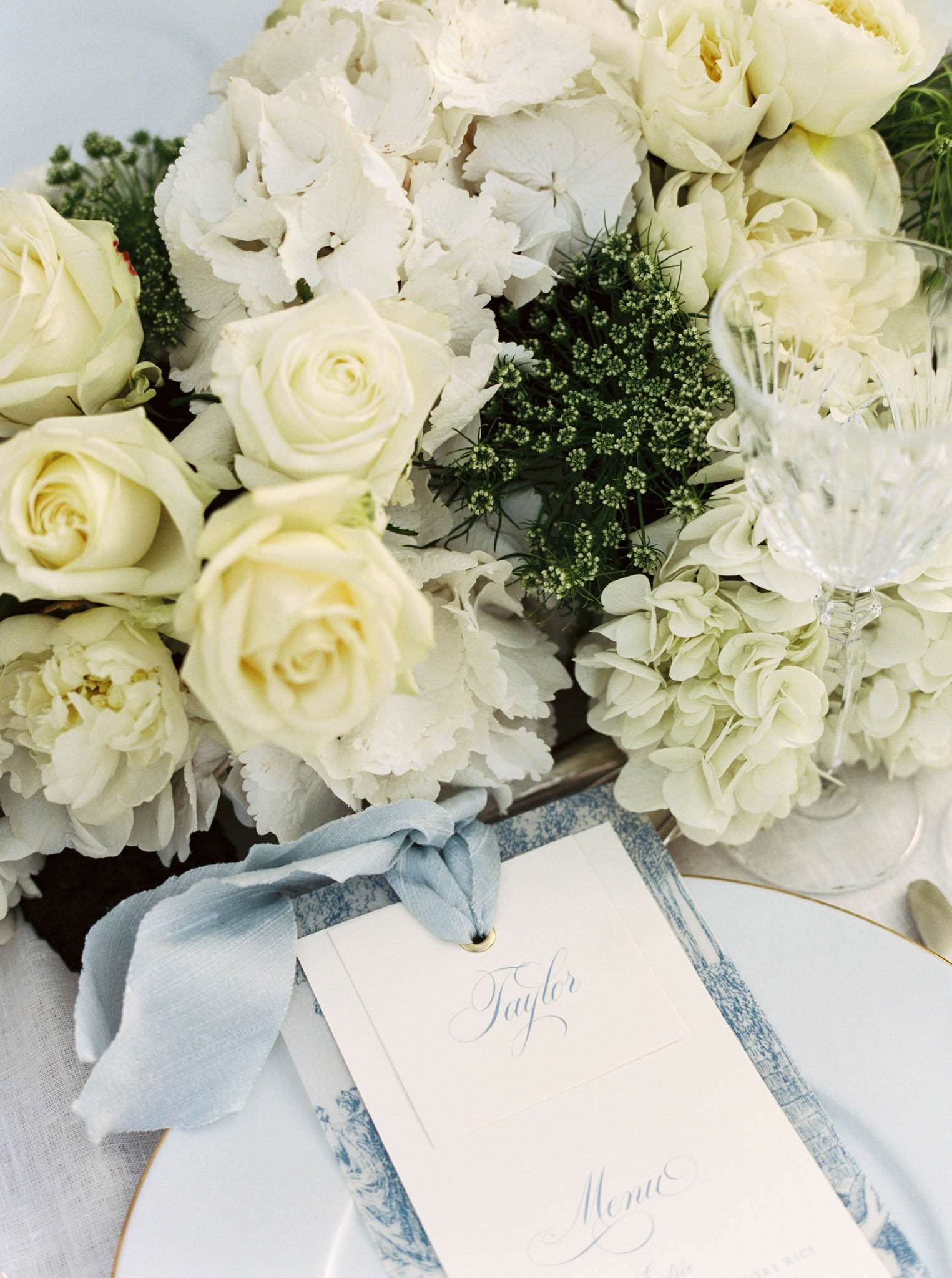 Reception place setting with gold-rimmed plate, blue toile charger, dusty blue menu card, and ivory roses