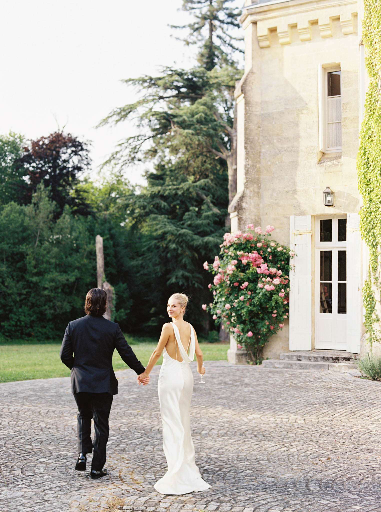 Couple walks hand-in-hand across cobblestone chateau courtyard, bride looking back holding champagne flute