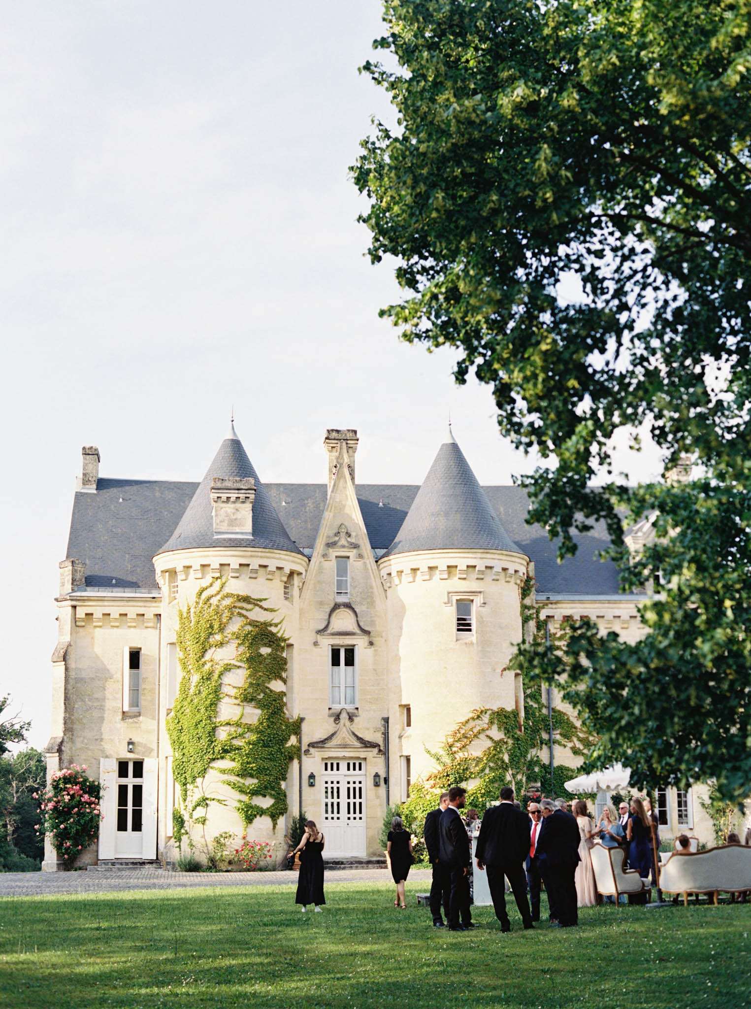 Guests gathered for cocktail hour on lawn of ivy-covered chateau with slate tower roofs in late afternoon light