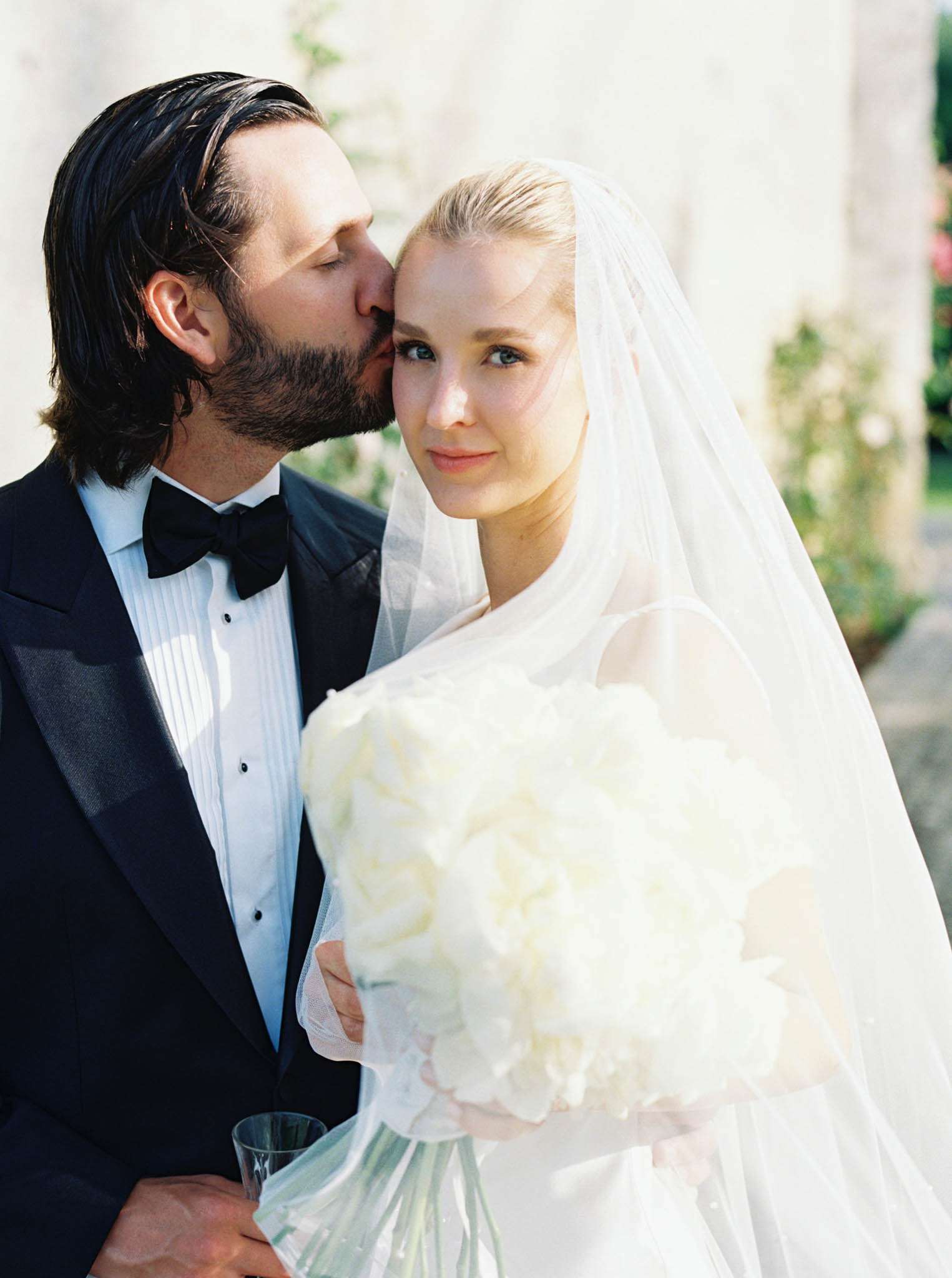 Groom in black tuxedo kissing bride on temple as she holds white peony bouquet against stone wall