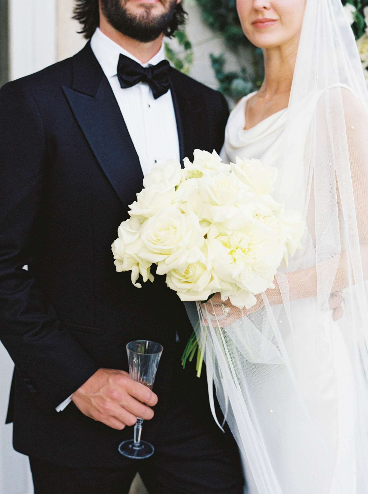 Cropped torso portrait of couple with white peony bouquet, pearl-trimmed veil, and champagne flute