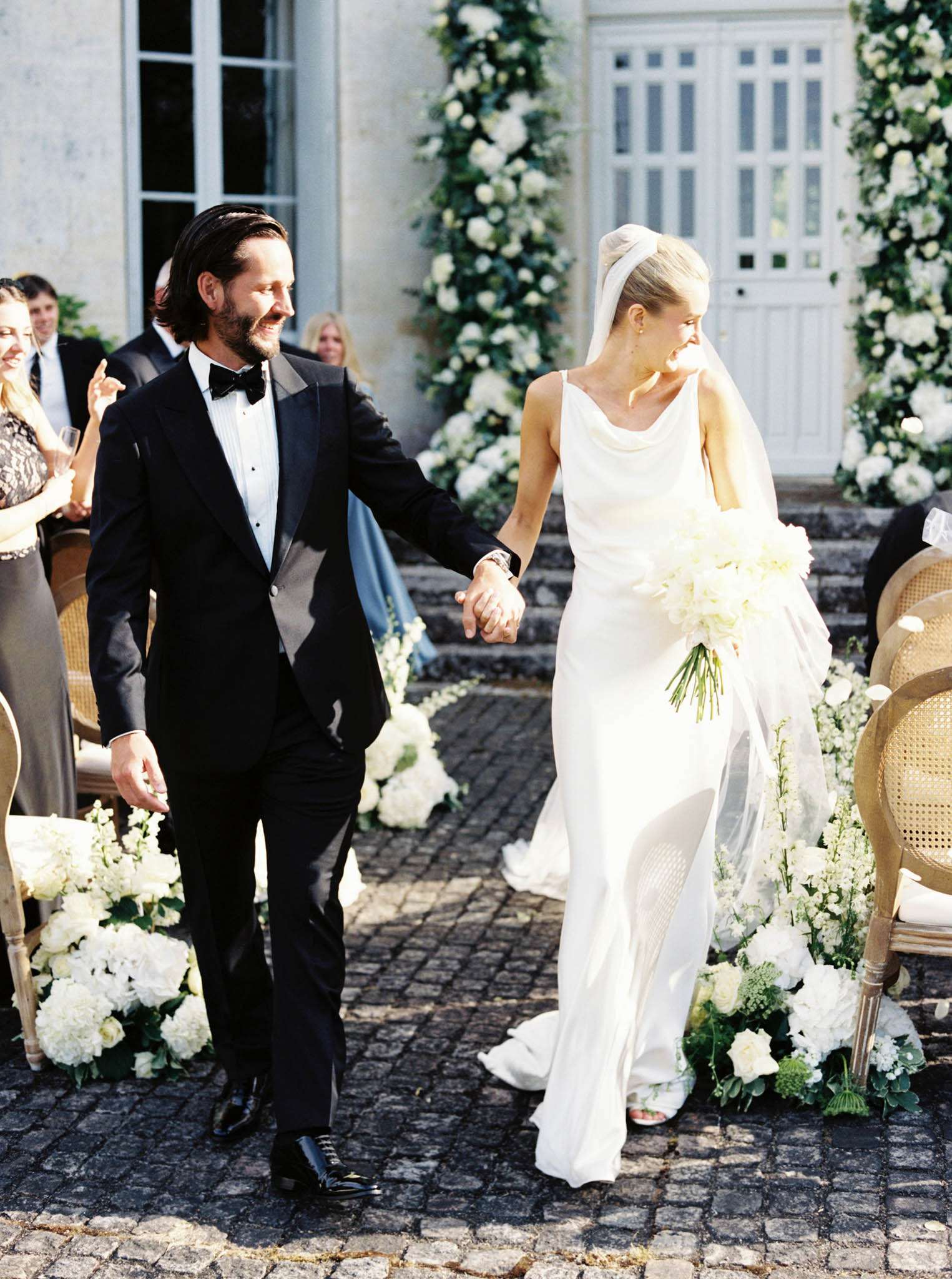 The bride and groom walk hand-in-hand back down the aisle following an outdoor ceremony, set on a cobblestone courtyard in front of a French château facade. The groom wears a black tuxedo with bow tie and the bride wears a sleek ivory cowl-neck slip gown with a long veil secured by a twisted fabric headband at the crown; she carries a bouquet of white peonies, hydrangeas, and greenery. The aisle is lined with low ground-level floral arrangements of white hydrangeas, roses, and stock flowers, and large white floral columns with climbing greenery frame the château's entrance doors in the background. Guests seated in cane-back chairs applaud on either side, and the overall decor palette is an all-white floral scheme with a modern classic styling. The shot is a medium portrait taken at eye level.