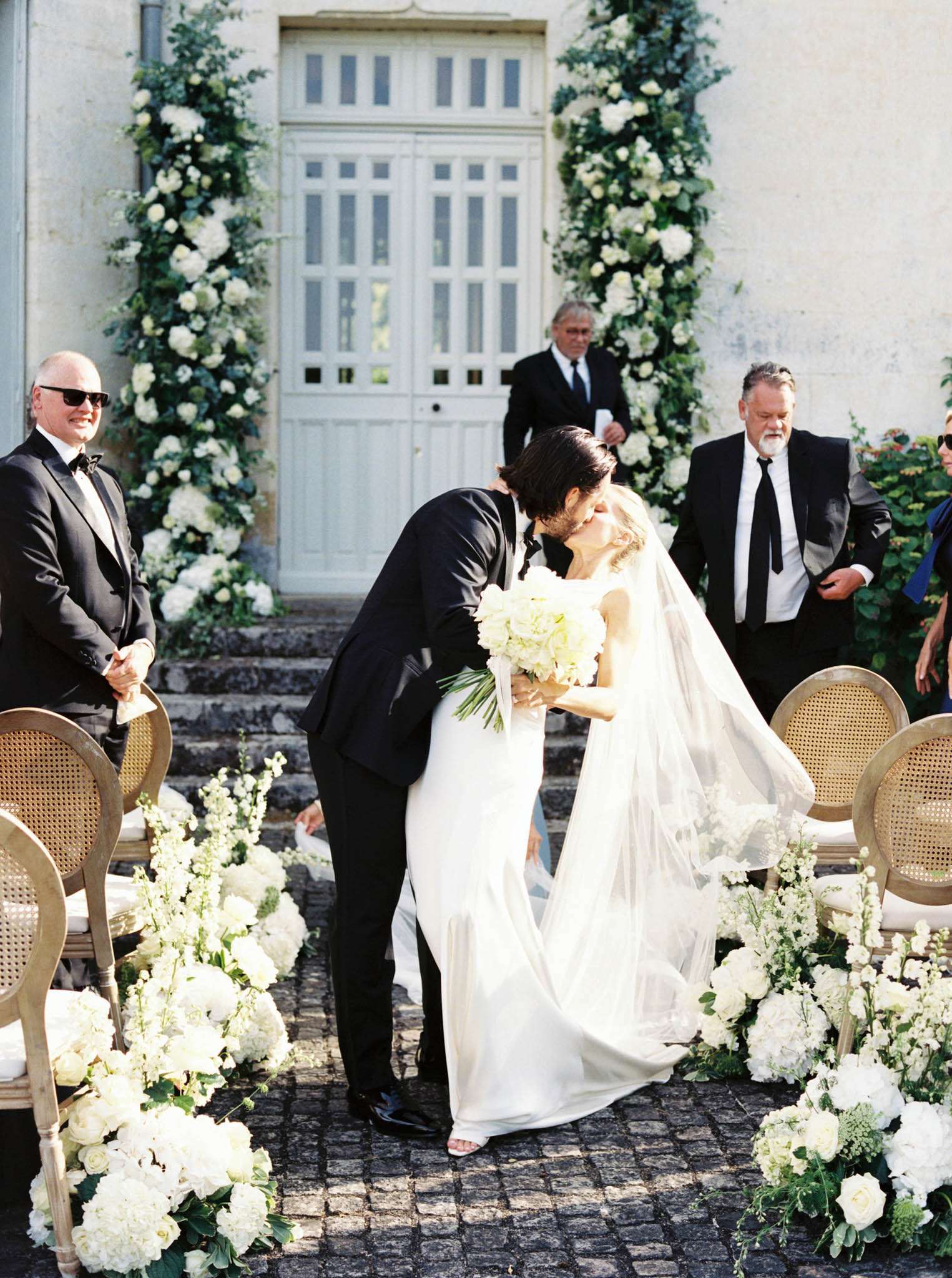 Couple shares first kiss at outdoor chateau ceremony with white hydrangea aisle and tall floral columns