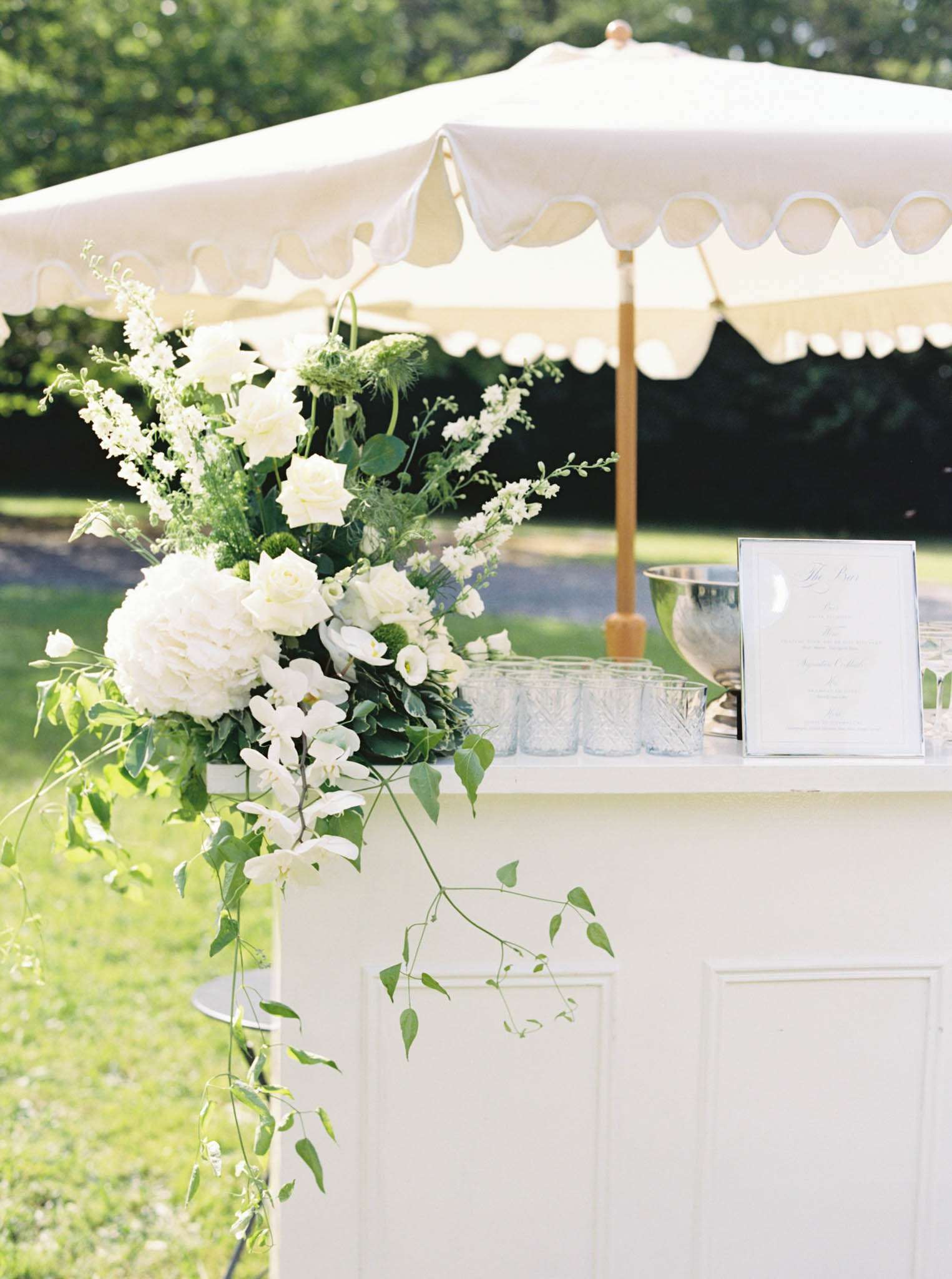 A detail shot of an outdoor cocktail hour bar station set up in a garden setting. The white painted bar counter is topped with a large floral arrangement featuring white peonies, white roses, white orchids, white lisianthus, and trailing green ivy and foliage that cascades down the front of the bar. On the bar surface, cut crystal glassware is lined up alongside a silver ice bucket and a silver-framed bar menu card with calligraphy script. A cream-colored market umbrella with a scalloped edge and wooden pole provides shade over the bar. The overall decor palette is white and green with a classic garden party aesthetic. Close-up detail composition with no people visible.