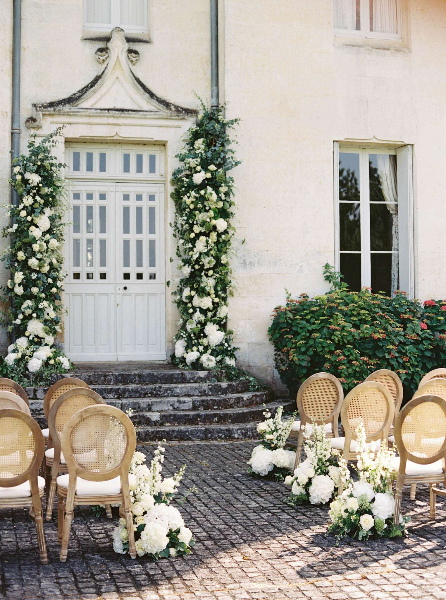 An outdoor wedding ceremony setup photographed in front of a French château façade, with no guests or couple present. The ceremony space is arranged on a cobblestone courtyard, with two rows of natural wood cane-back Louis XVI-style chairs with cream cushions forming an aisle. White hydrangeas, white delphiniums, and green foliage are arranged in low clusters along the aisle floor. The château's white painted double door serves as the ceremony backdrop, flanked on both sides by tall, lush floral columns featuring white hydrangeas, white roses, and trailing greenery including eucalyptus and ivy. The overall floral and decor palette is white and green, with a classic French formal styling. Wide shot capturing the full ceremony space and building façade. Potential venue feature image.