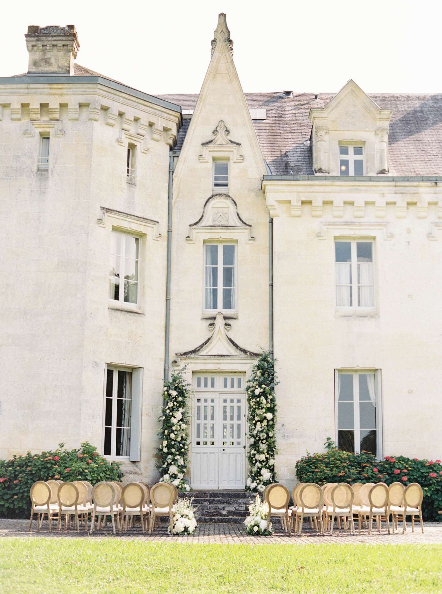 An outdoor wedding ceremony setup photographed in a wide shot directly facing the facade of a French château. Two rows of gold-framed Louis XVI-style chairs with ivory cushions are arranged in an aisle formation on a cobblestone courtyard in front of the château's central entrance door. The pale grey double door is framed by lush floral installations of white blooms — including what appear to be white roses and hydrangeas — combined with trailing greenery climbing either side of the doorframe. Small white floral clusters are also placed at ground level flanking the base of the steps. The château facade is cream-toned limestone with Gothic-influenced architectural detailing, including a pointed dormer above the central entrance. Potential venue feature image.