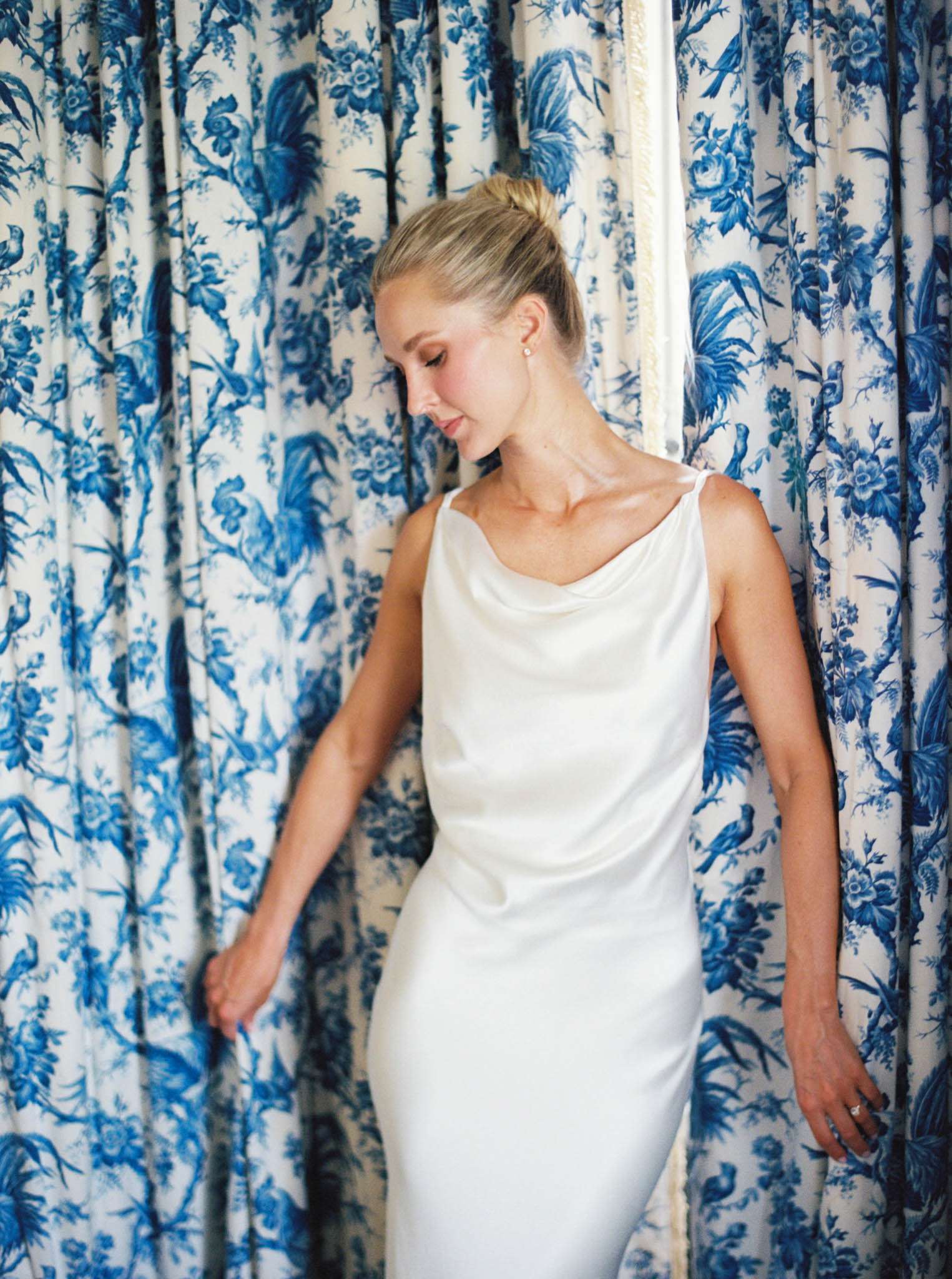 Bride in ivory satin slip dress standing in front of blue and white toile de Jouy curtains