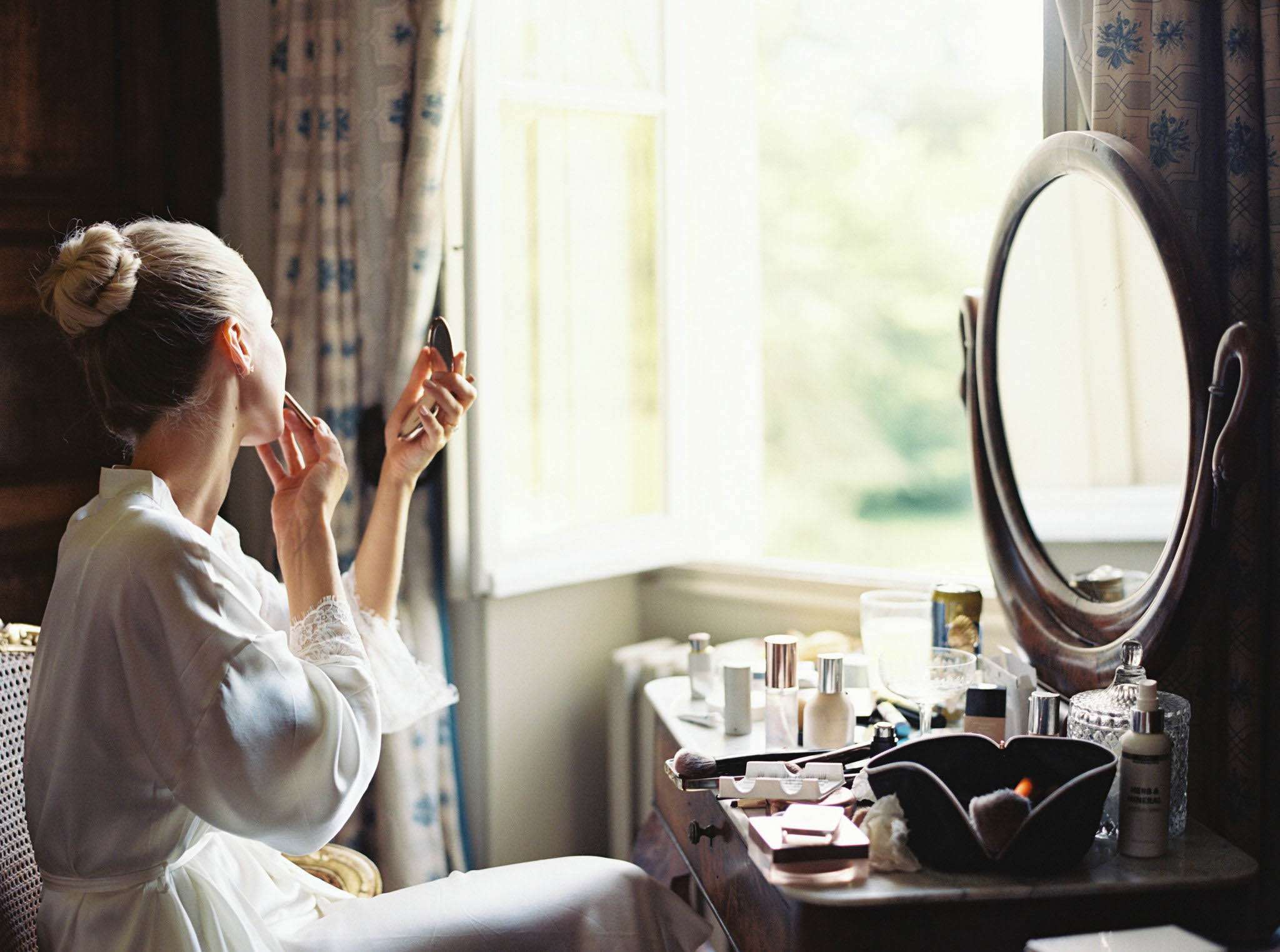 Bride in white robe applying makeup at antique vanity beside blue floral curtains