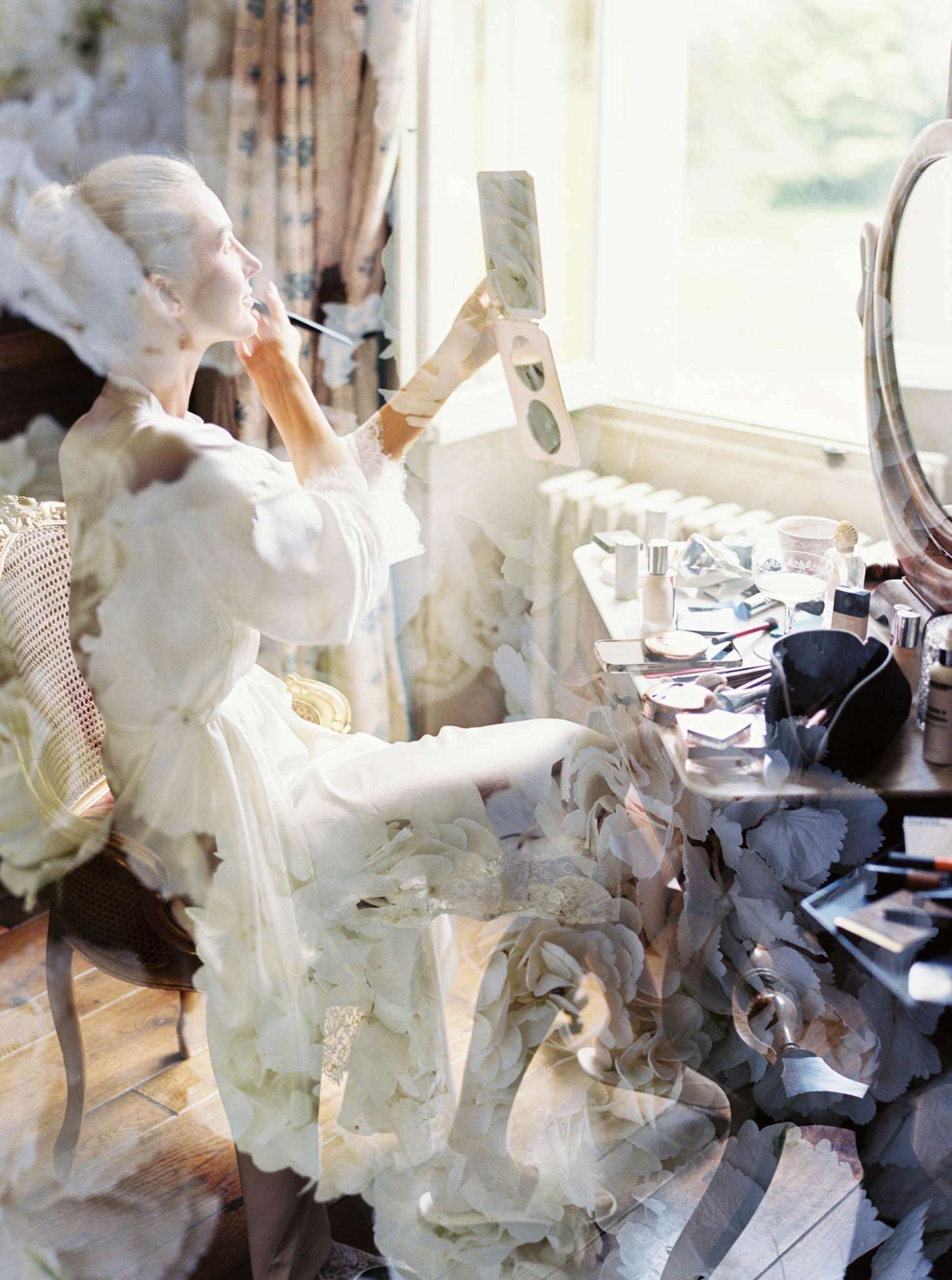 A double-exposure or in-camera multiple-exposure image taken during bridal preparations, showing a bride seated at a dressing table applying makeup while holding a small compact mirror. She is wearing a white lace-trimmed bridal robe and has her hair pinned up with a white hair accessory. The dressing table is covered with an array of makeup products, brushes, perfume bottles, and beauty tools, with a large round vanity mirror visible to the right. A floral-patterned curtain and bright natural window light frame the scene. The double-exposure overlays a close-up of cream-white hydrangea blooms semi-transparently across the entire composition, blending the bride and the florals into a single layered image. The setting appears to be a classically decorated indoor room with wooden floors and cane-back chair, suggesting a château or manor house getting-ready space. The overall shot is a creative medium portrait with a soft, high-key, film-grain quality.